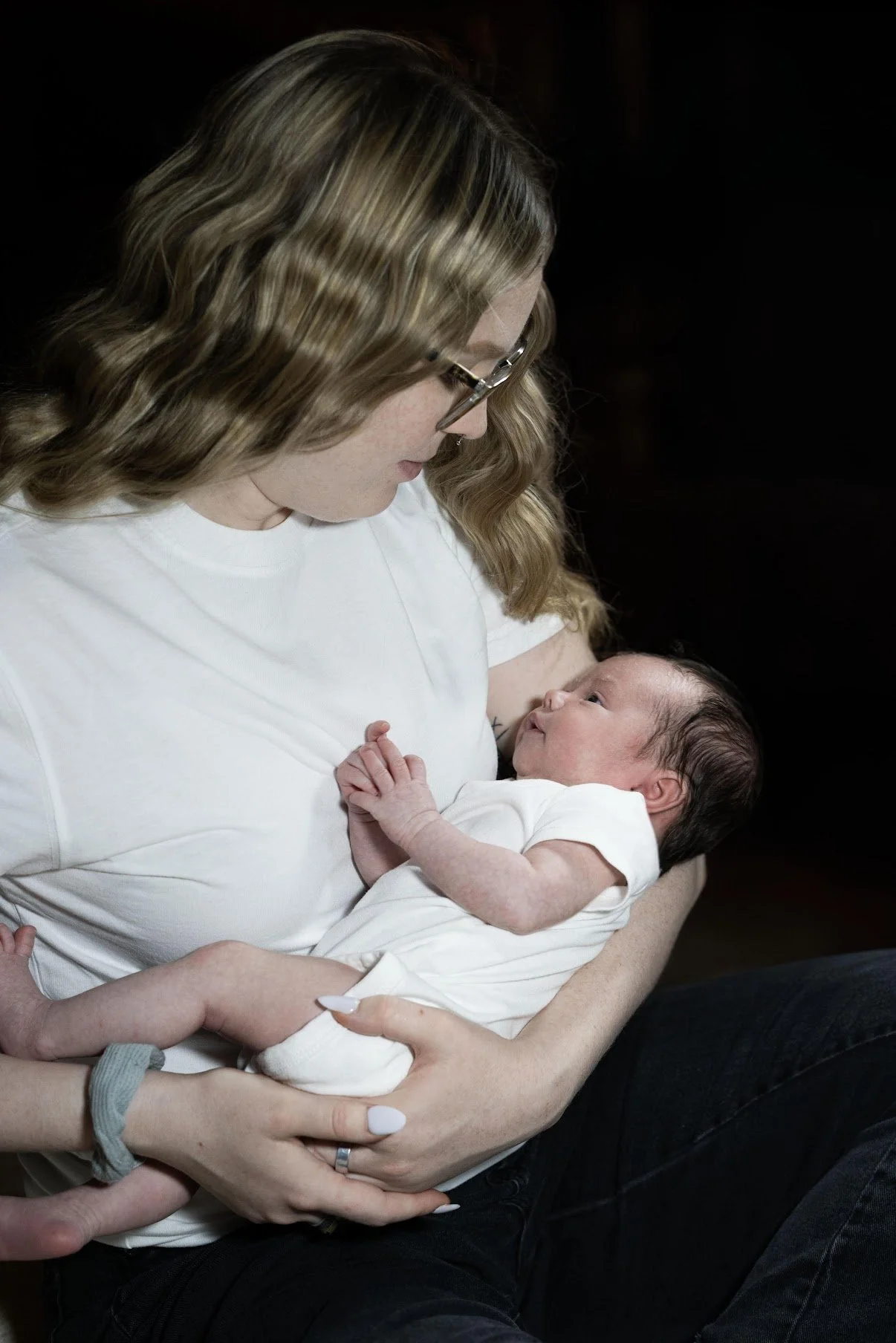 A woman with wavy blonde hair and glasses holds a newborn baby close to her chest, looking down at the baby, who is dressed in a white outfit and gazing back at her.