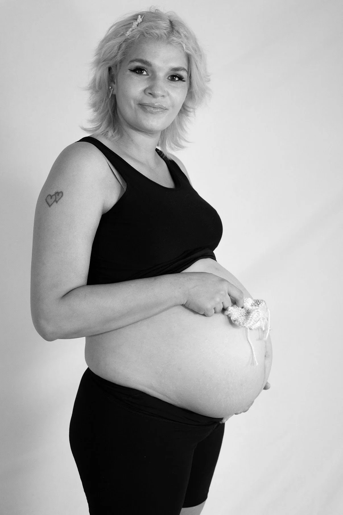 A pregnant woman with blonde hair, wearing a black sleeveless top and black pants, holding her pregnant belly with a cloth and smiling.