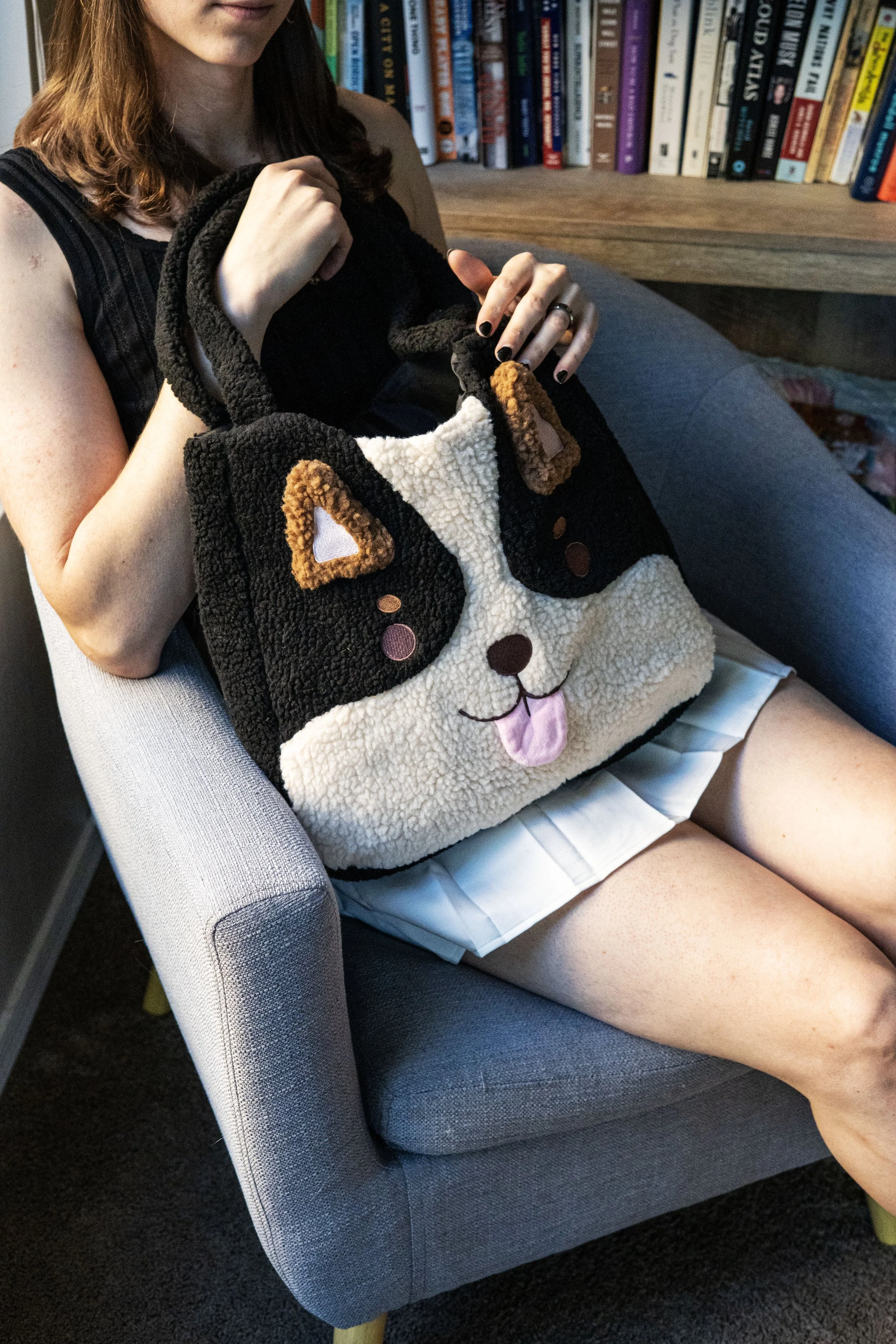 A woman sitting in a light gray armchair holding a black and white plush bag shaped like a dog's face. The woman has shoulder-length brown hair, is wearing a sleeveless top, and a white skirt. Behind her, there is a wooden bookshelf filled with color