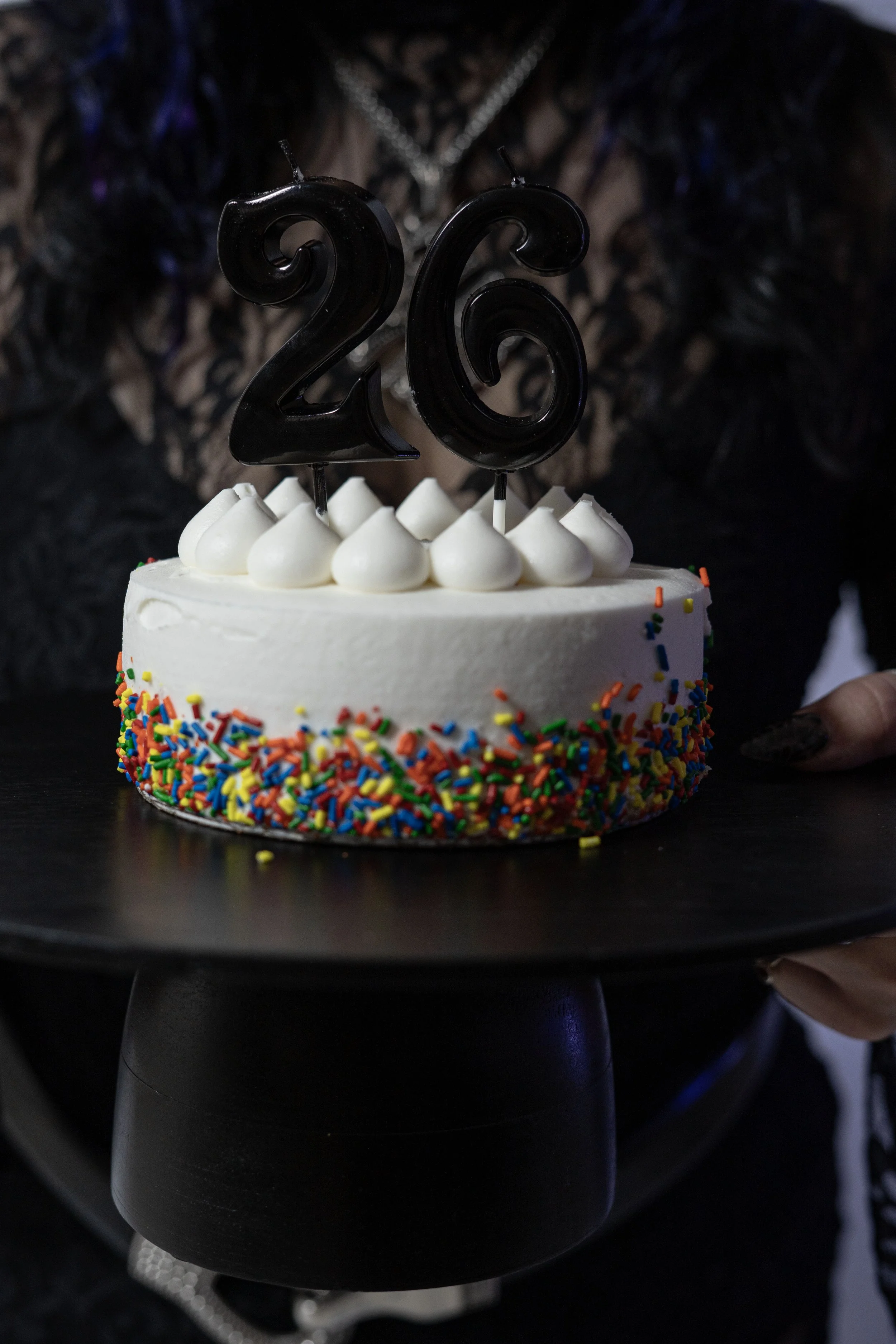 A birthday cake decorated with white frosting, colorful sprinkles on the sides, and white dollops on top with black number candles '26' on top.