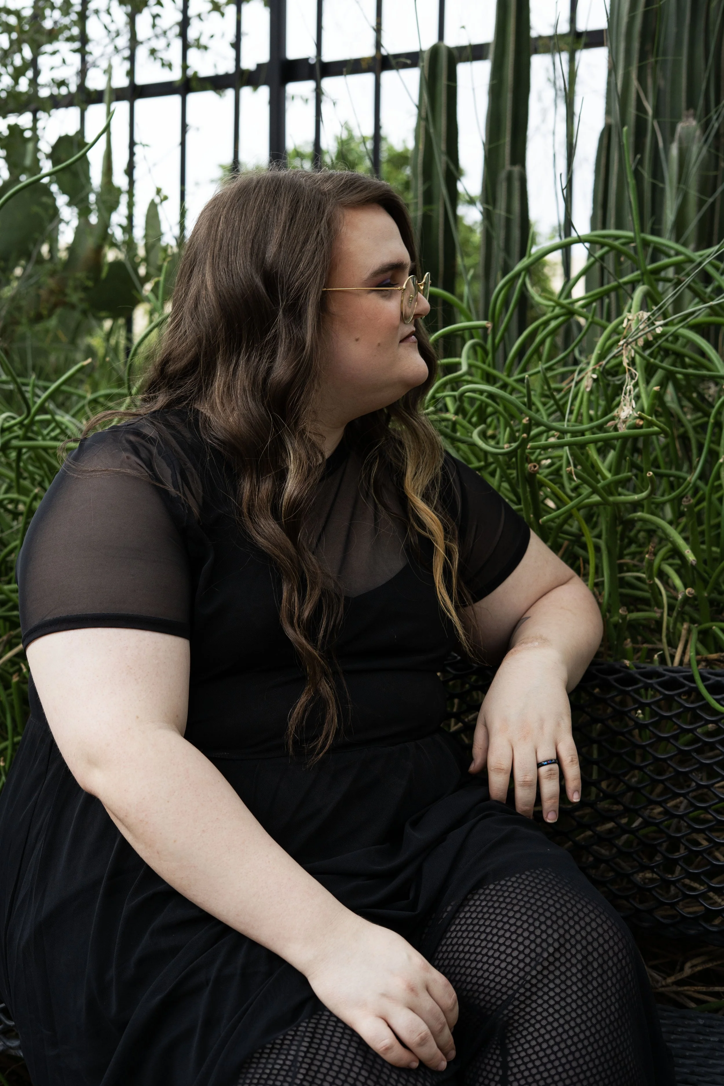 A woman with long brown wavy hair, glasses, wearing a black dress, sits on a bench next to green plants with vine-like stems, inside a greenhouse with glass panels.