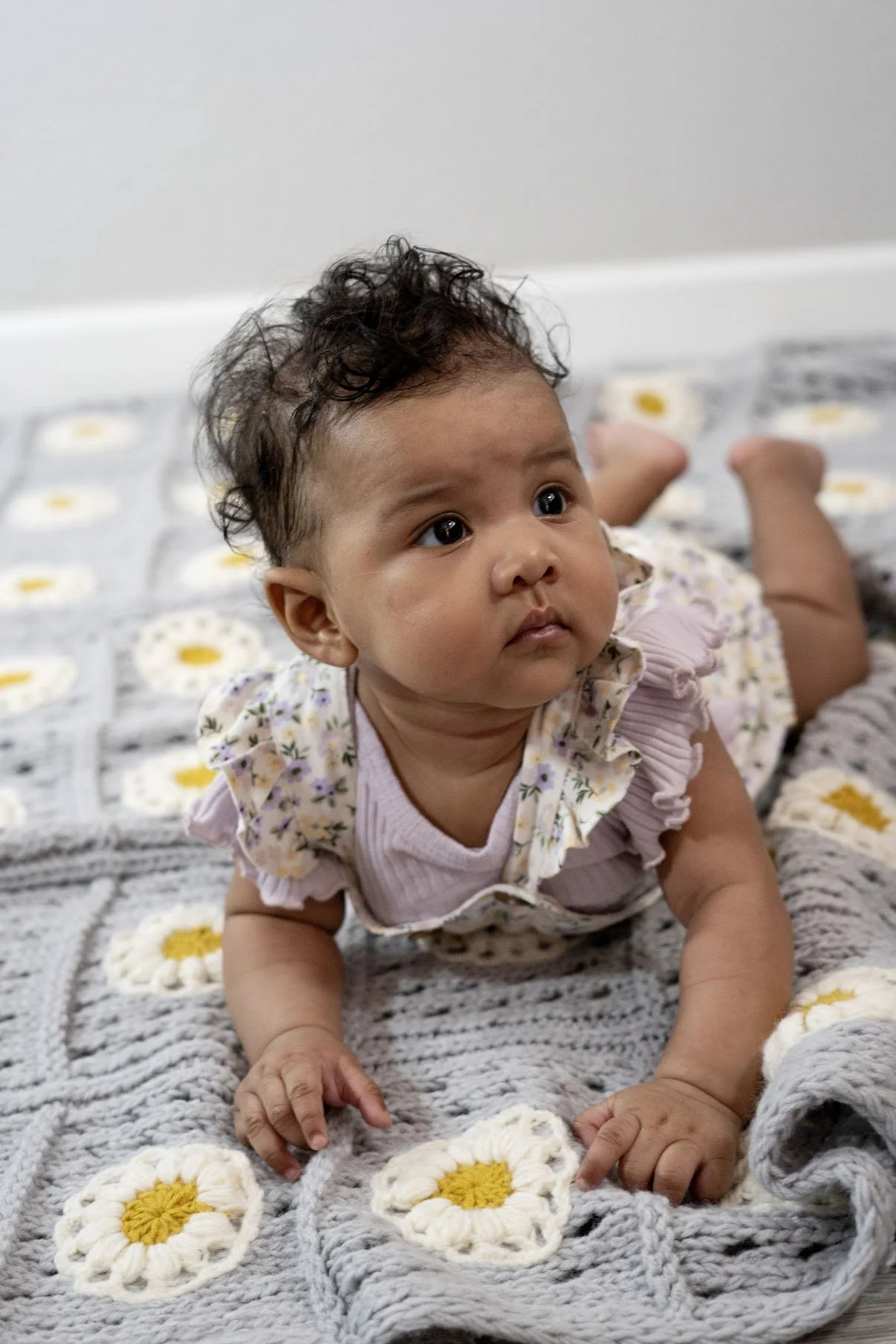 A baby girl with curly dark hair lying on her stomach on a gray blanket with white and yellow daisy patterns.
