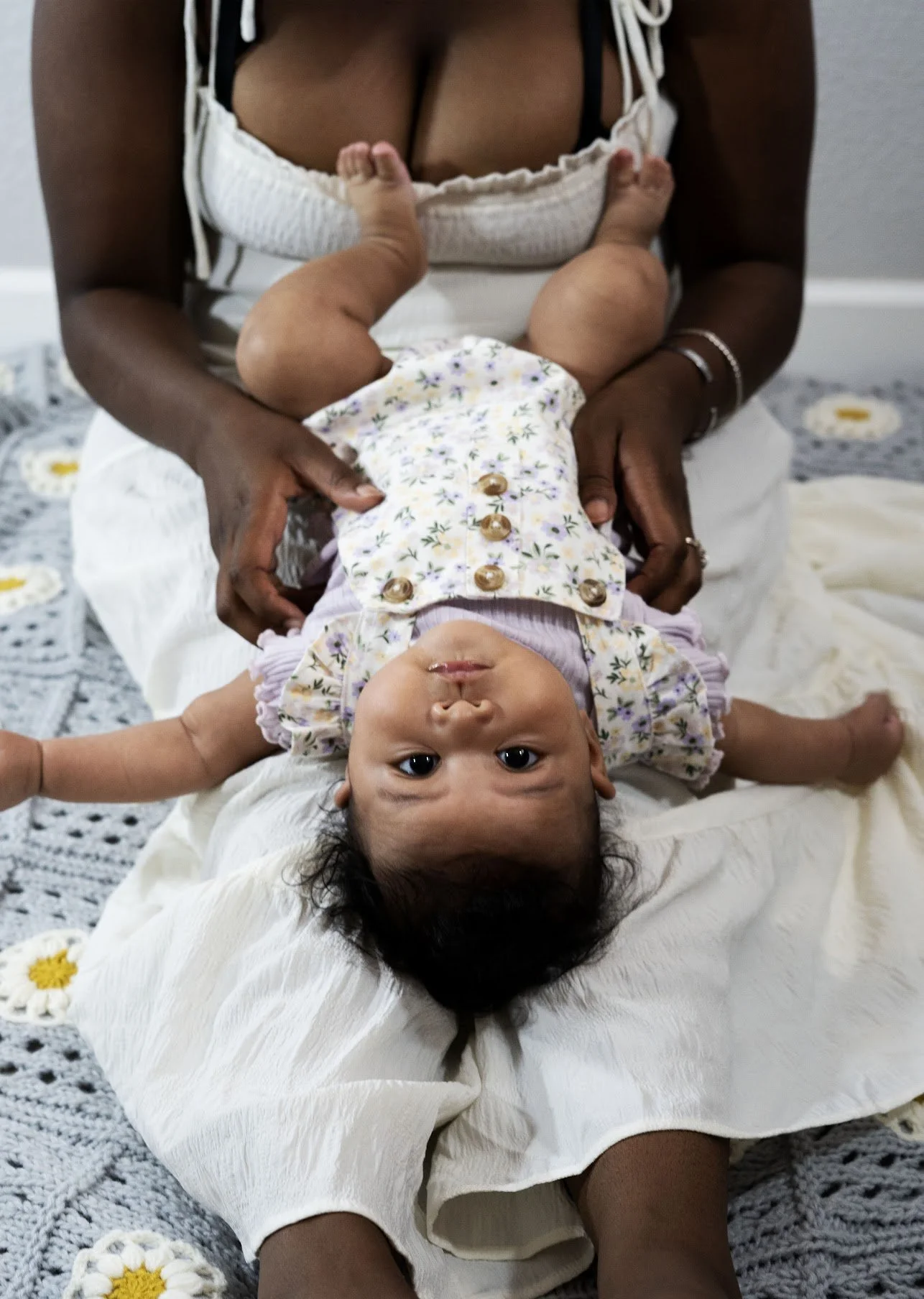 An adult woman holding a baby upside down on her lap, with the baby's head near the woman's abdomen and feet pointing upward. The woman is sitting on a gray crocheted blanket with white and yellow flowers, wearing a white dress. The baby is dressed i