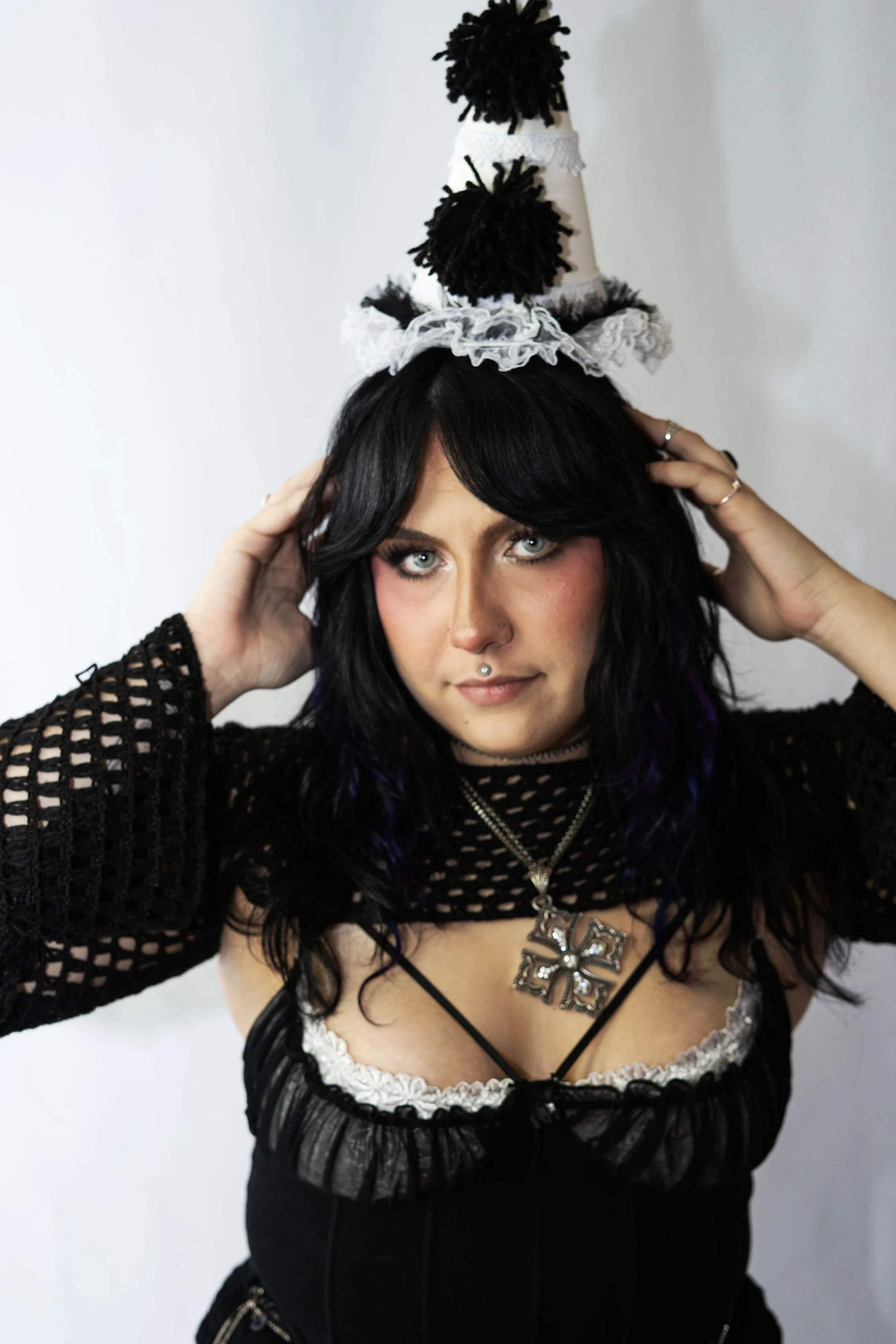 Woman with black hair and blue eyes wearing a black mesh top, layered jewelry, and a white and black party hat with pom-poms.