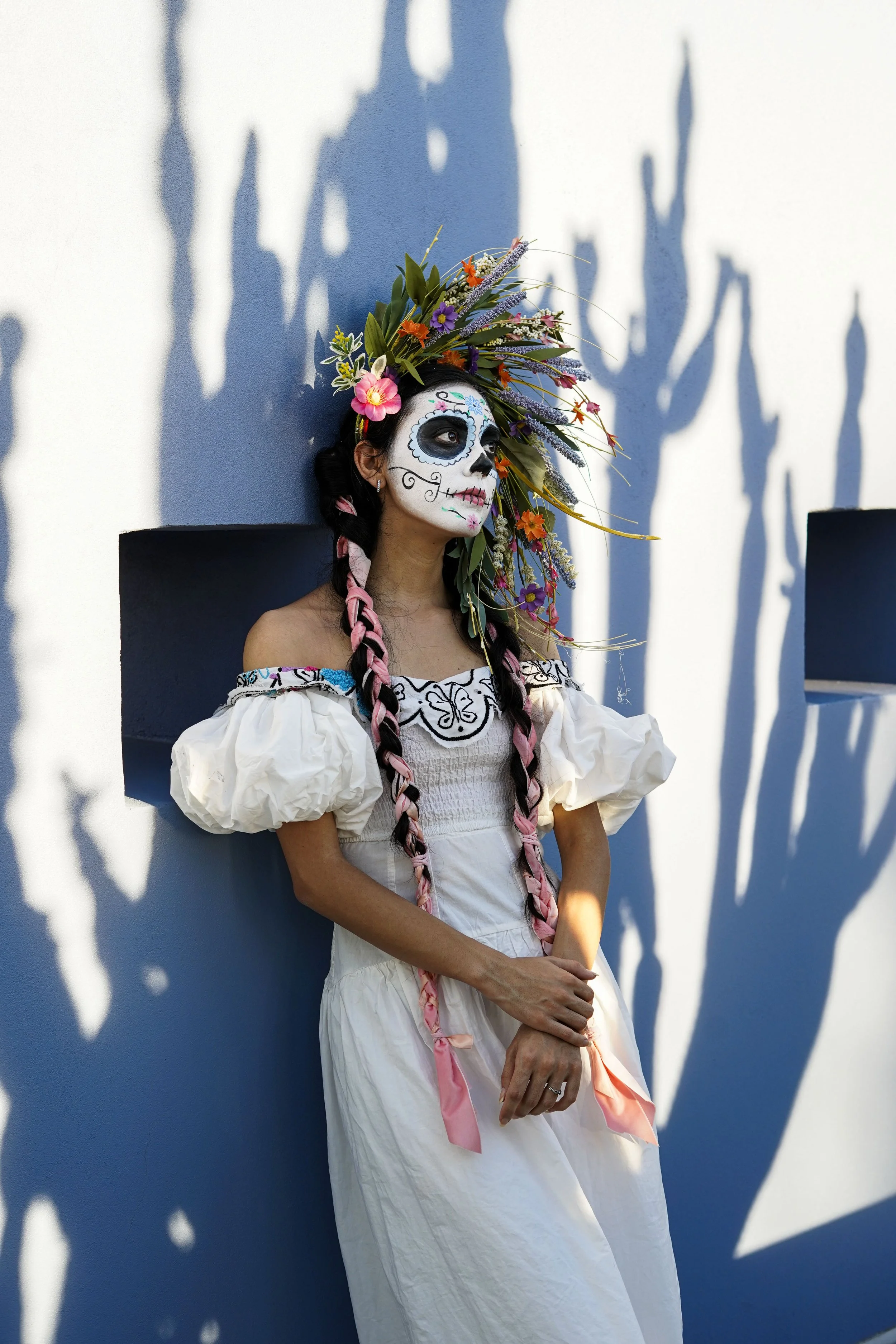 Woman dressed in traditional Mexican attire with sugar skull makeup and a floral headdress, standing against a white wall with shadows.