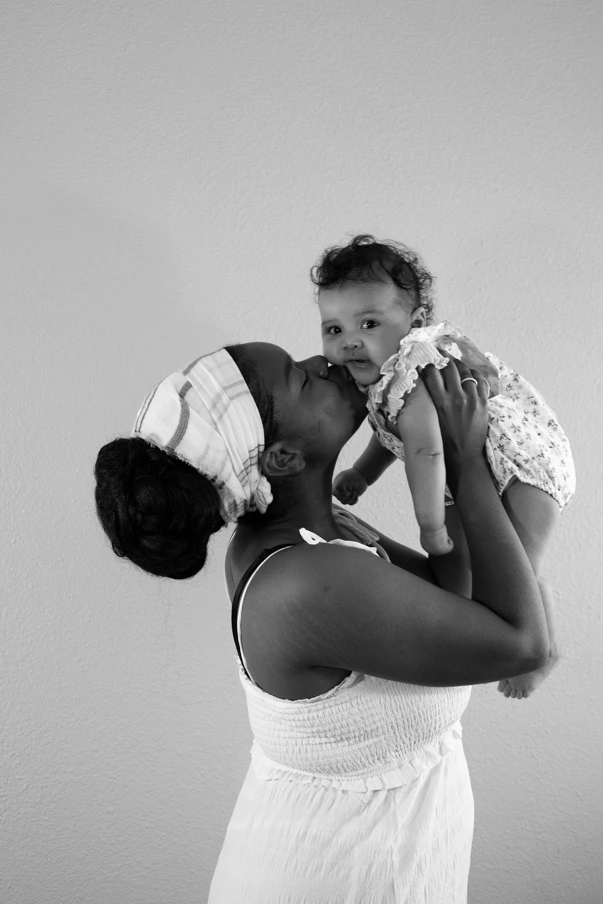 A woman holding a toddler girl in her arms, kissing her on the cheek, with both smiling. The photo is in black and white, with a plain wall in the background.