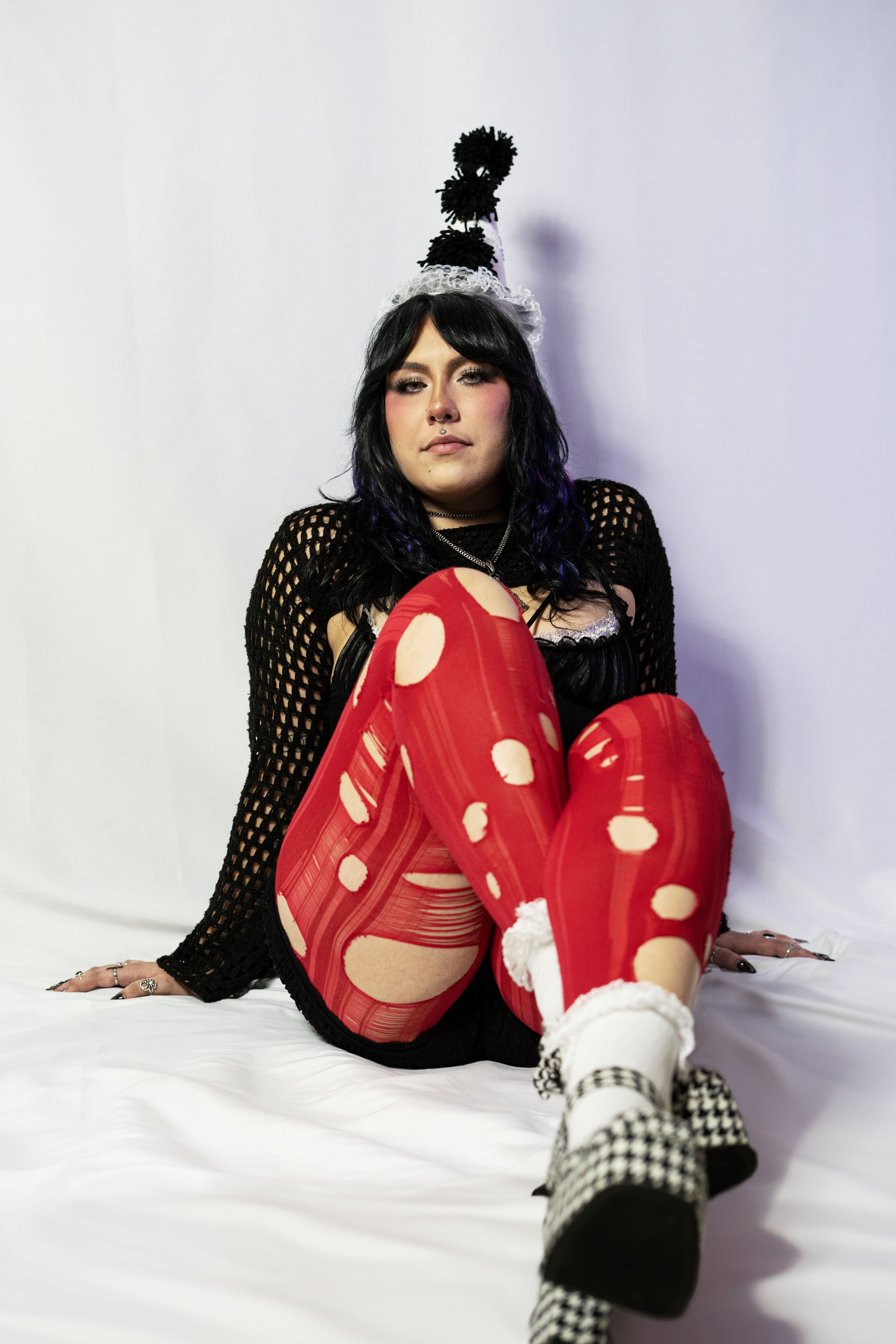 A woman sitting on a white surface wearing red distressed tights, black clothing, and checkered boots, with a black and white party hat on her head, against a plain white background.