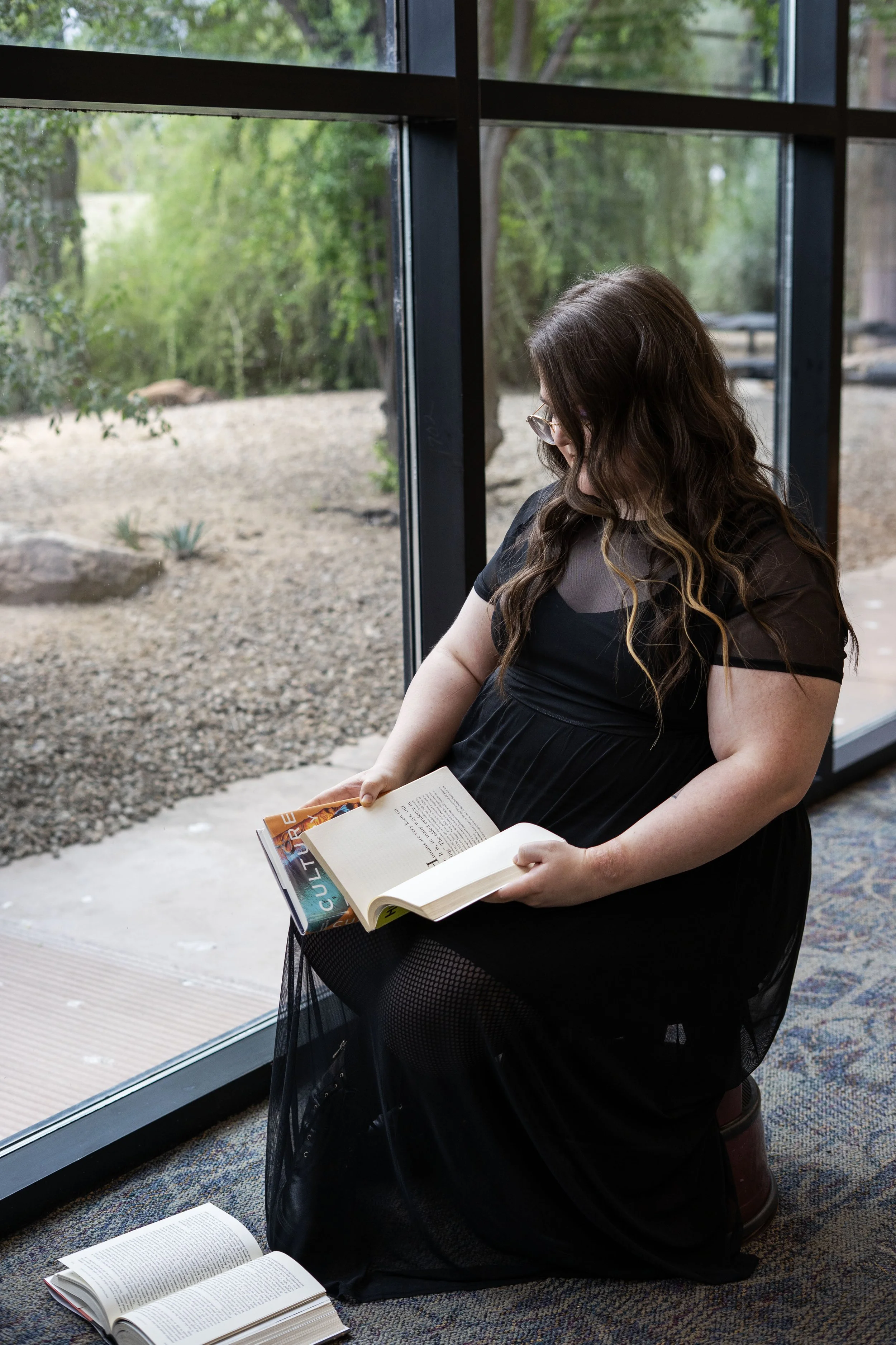 A woman with long wavy hair, wearing glasses and a black dress, sitting on a small stool by a large window, reading a book.