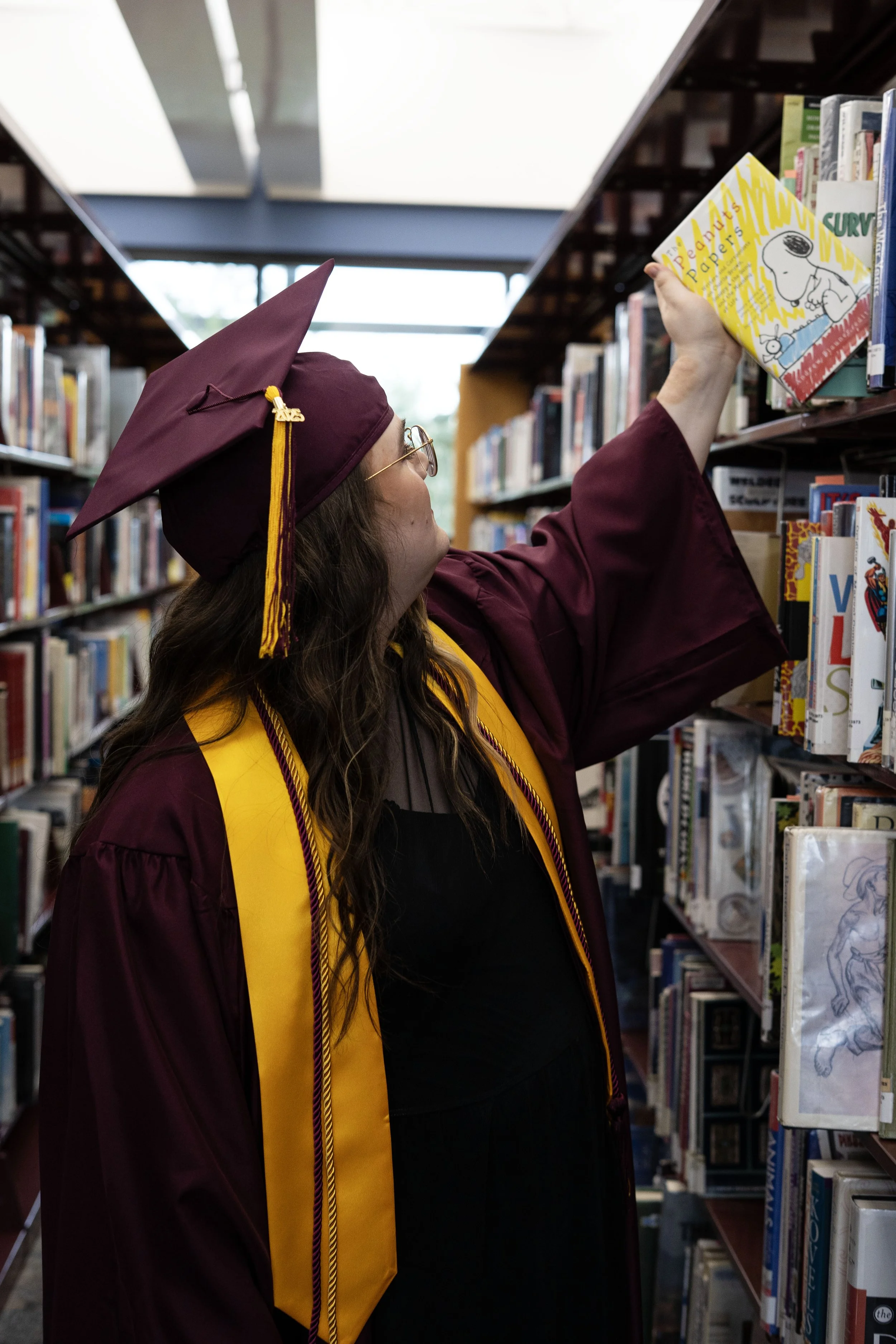 Graduate in maroon cap and gown reaching for a book on library shelf.