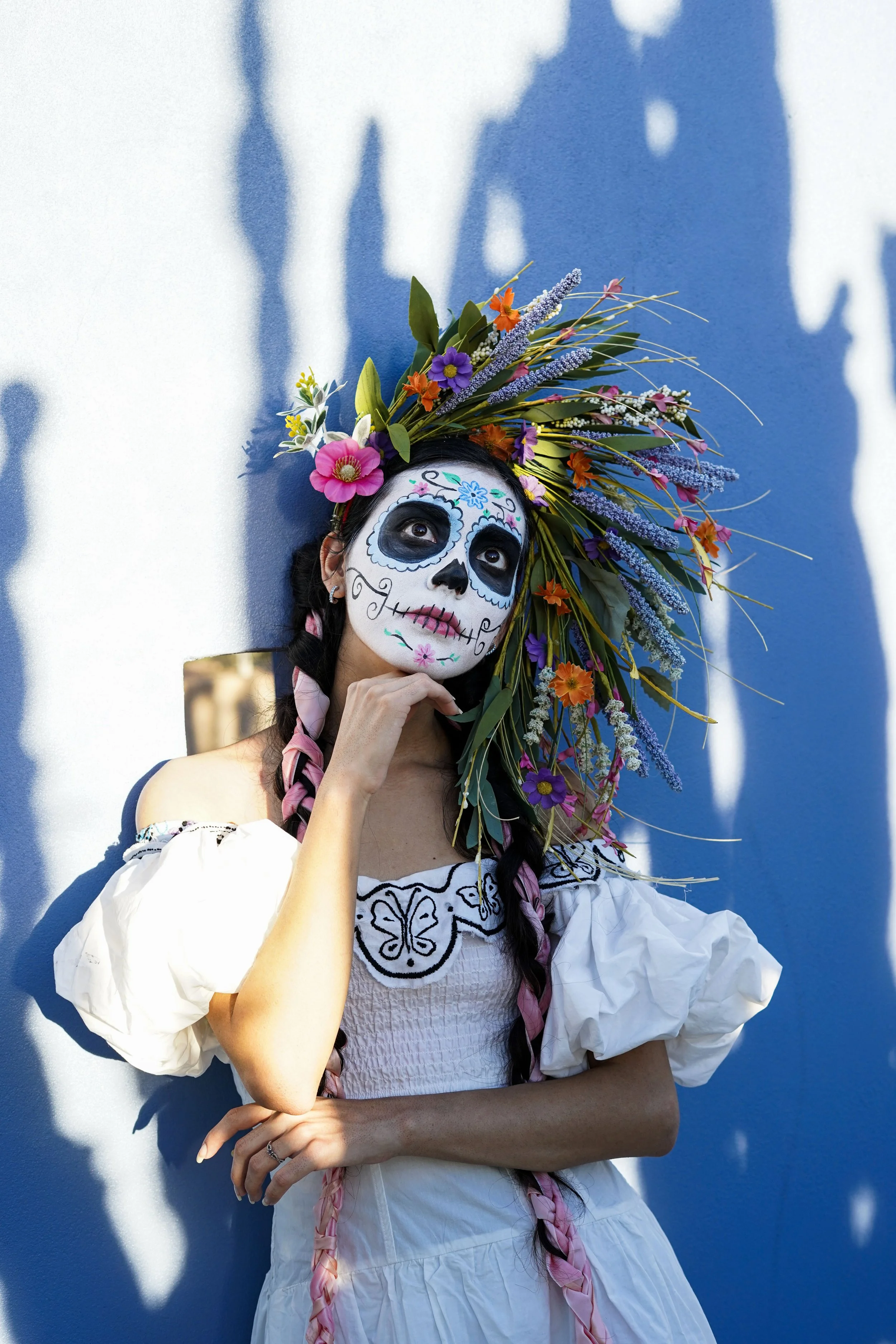 A woman with sugar skull makeup and colorful flower crown on her head standing against a blue wall with shadows.
