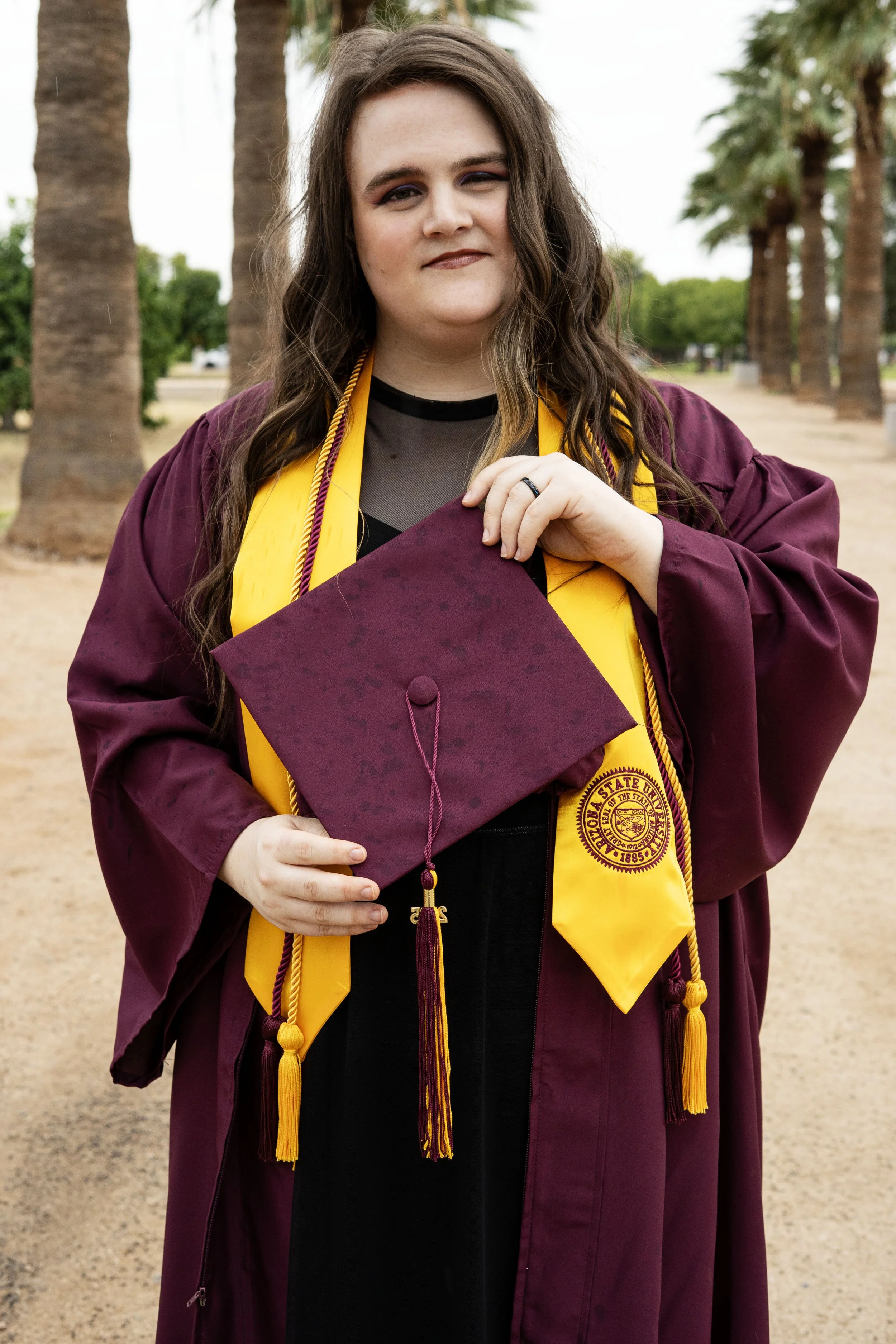 A woman wearing a maroon graduation gown and yellow stole holding a maroon mortarboard cap, standing outdoors among palm trees.