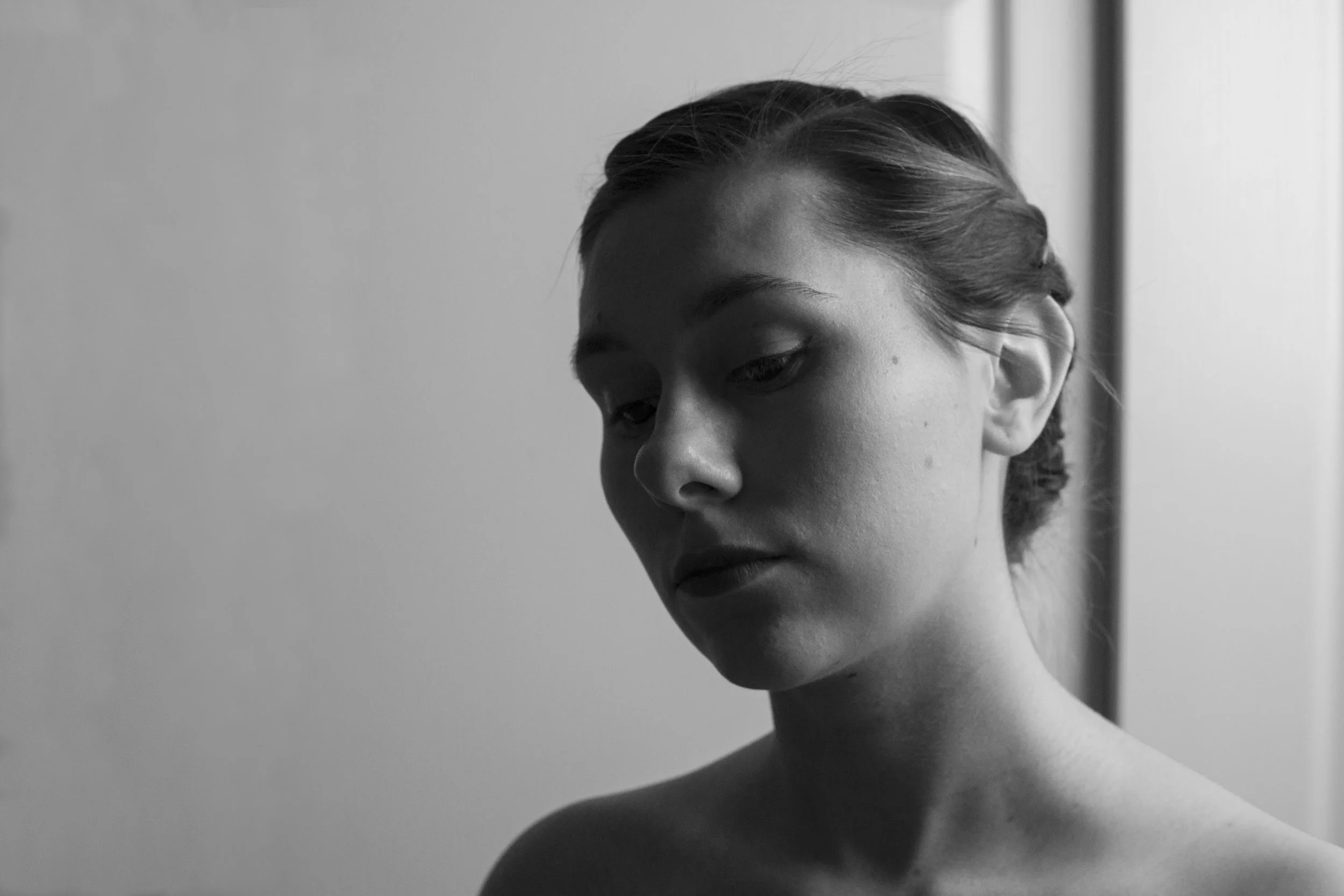 Black and white photo of a woman with her hair styled back, looking down with a serious expression, near a window.