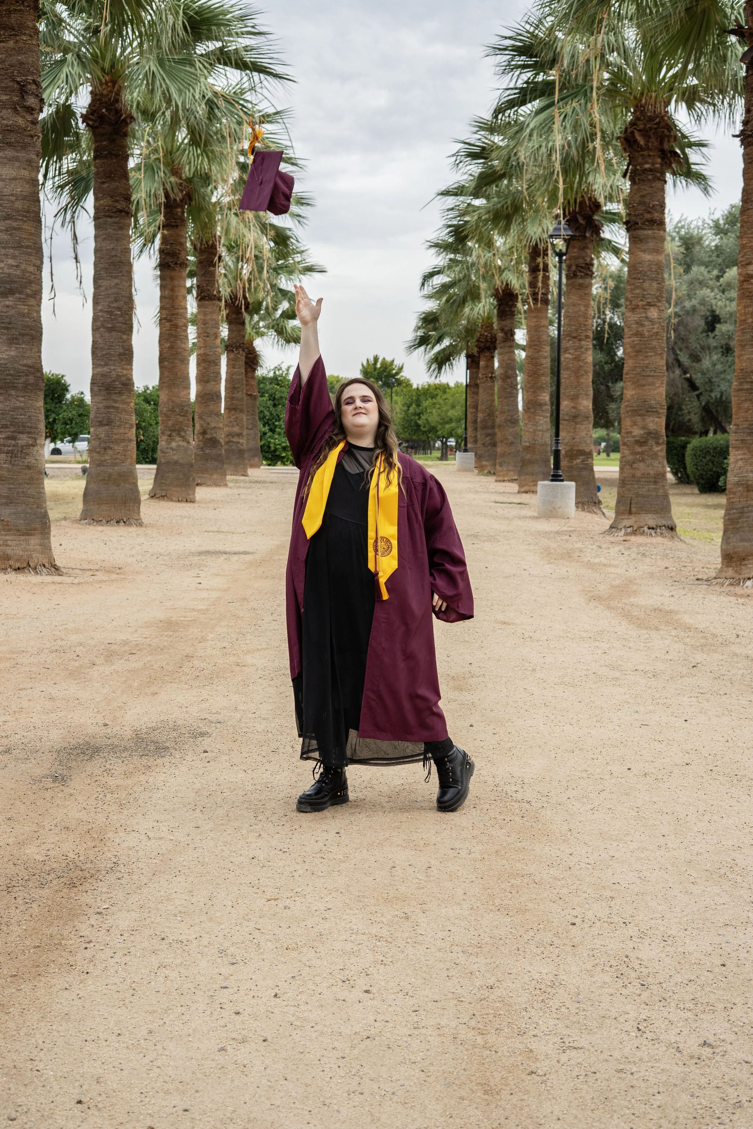 A young woman in a graduation gown and cap tossing her cap in the air in a park lined with palm trees.