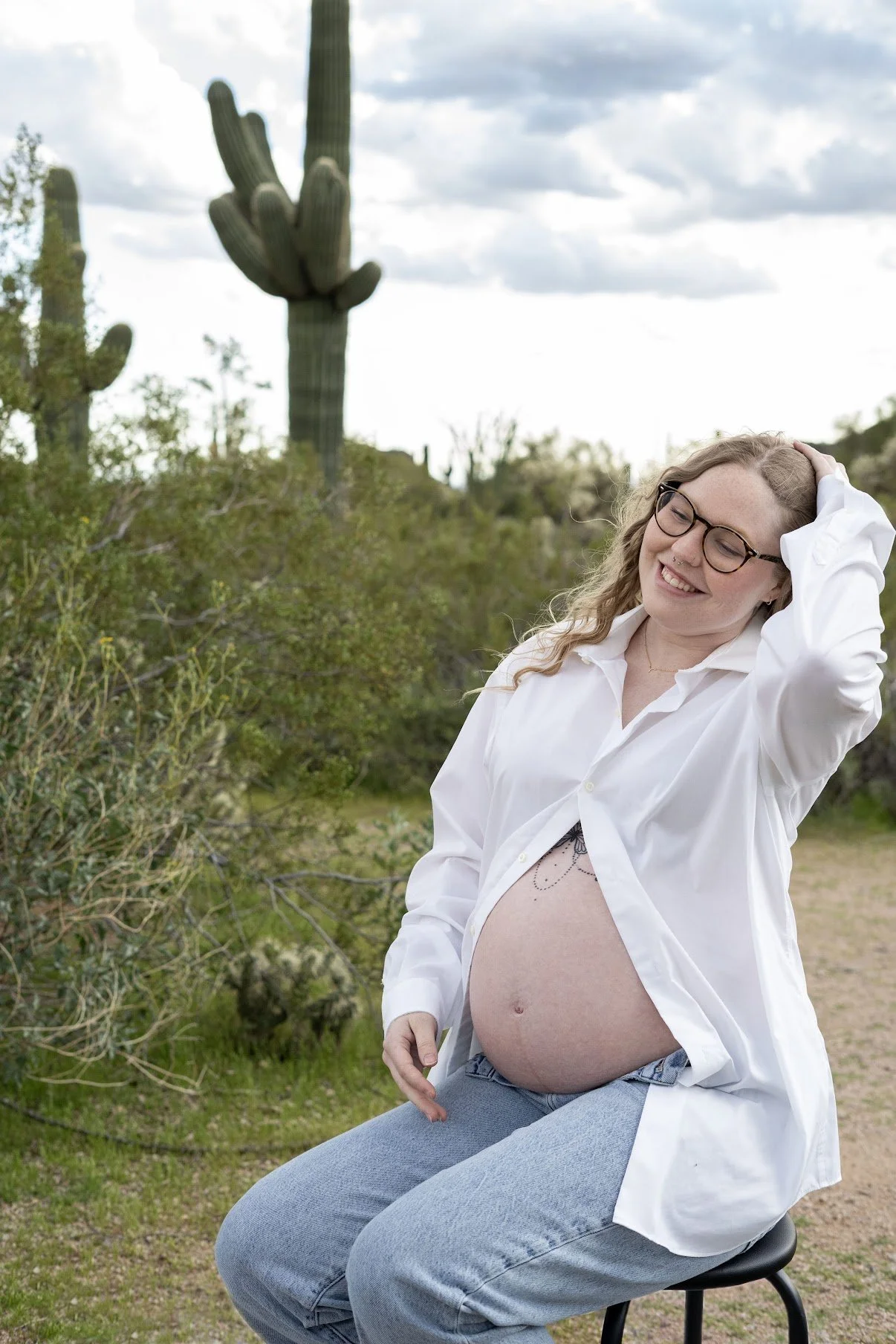 A pregnant woman wearing glasses and a white shirt sitting on a stool outdoors, smiling with her hand on her head, in a desert landscape with a cactus in the background.