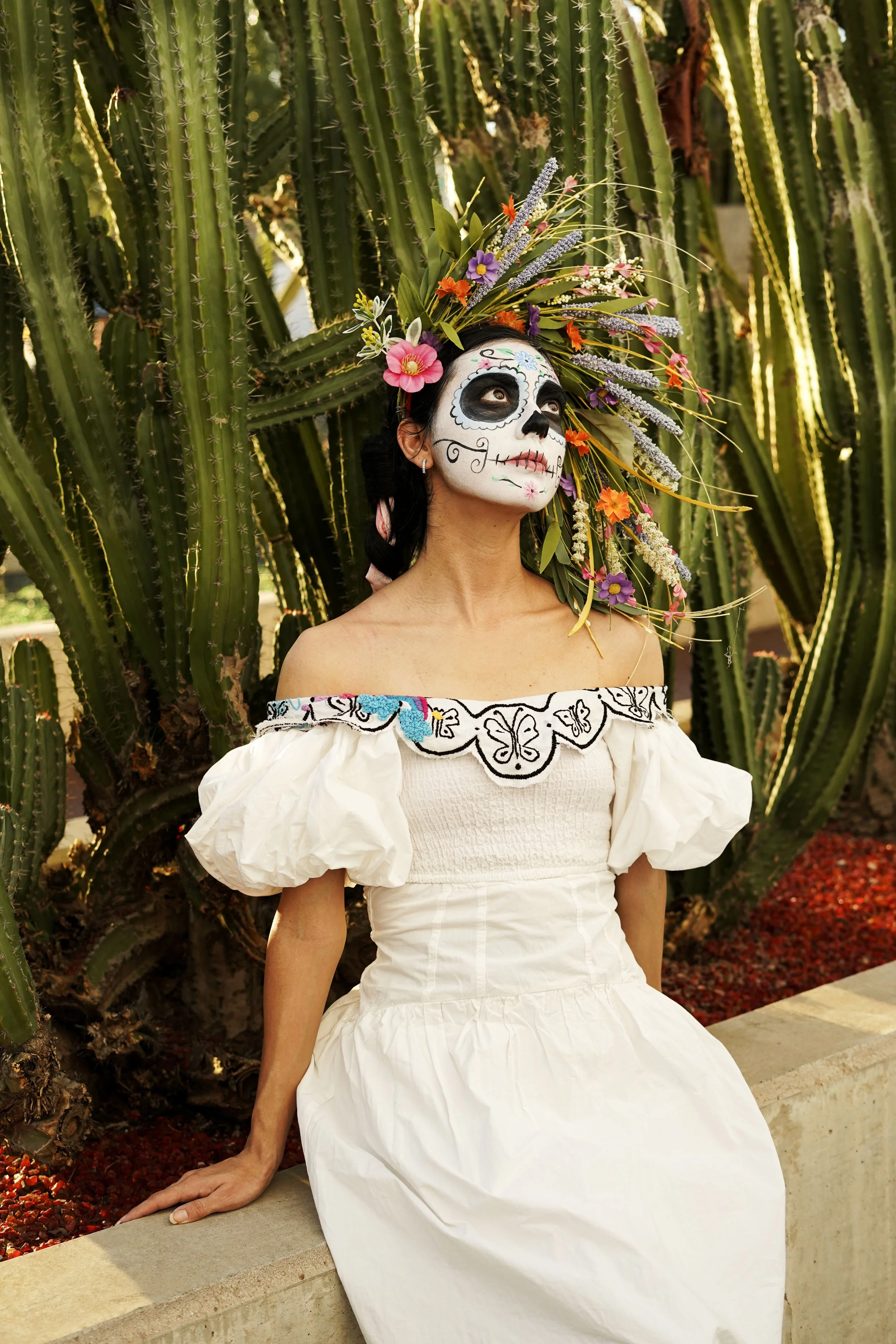 A woman dressed in traditional Mexican attire with sugar skull face paint, sitting on a concrete block in front of a large cactus, wearing a white off-the-shoulder dress and a colorful floral headdress.