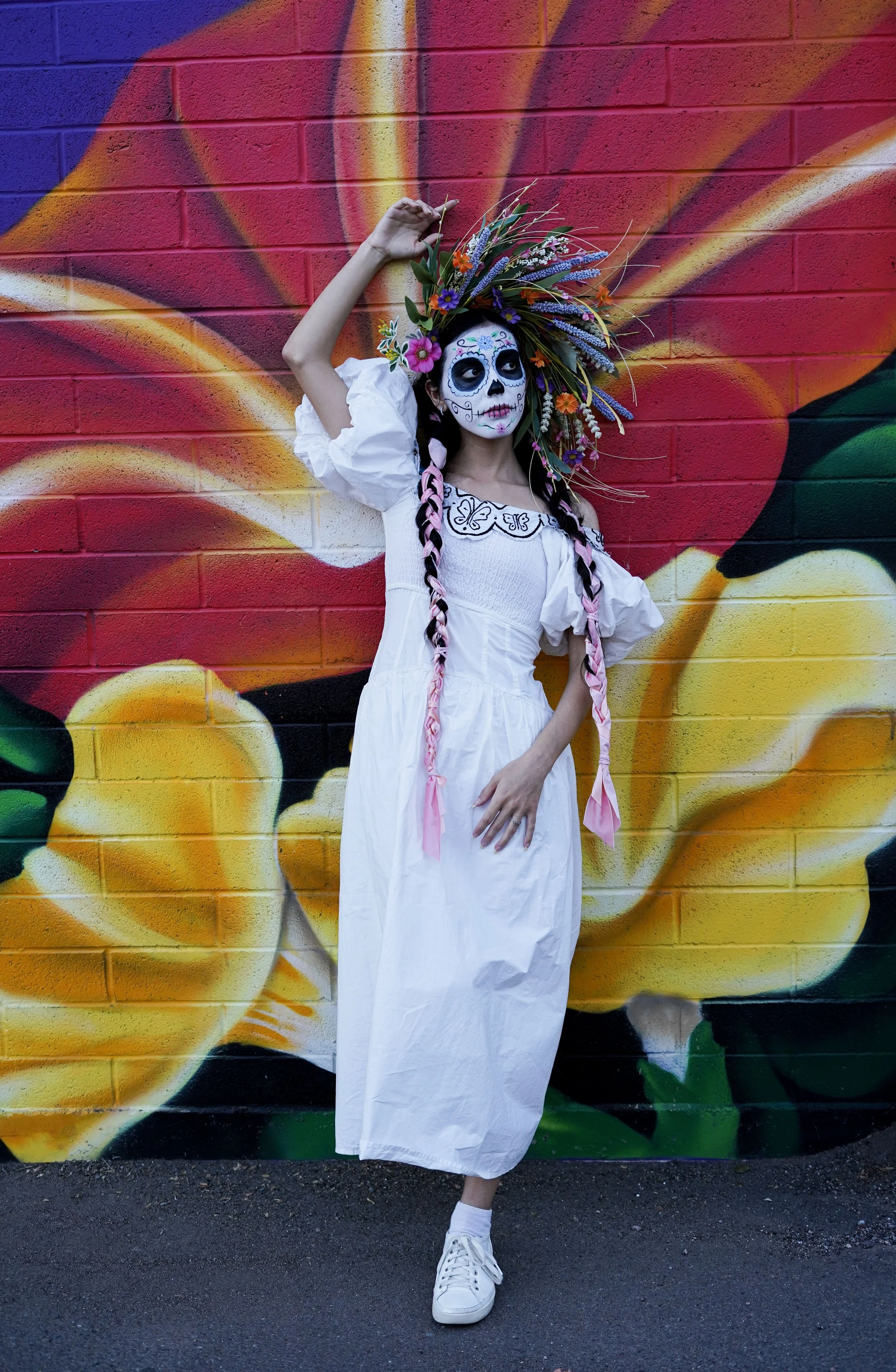 A young woman dressed as a Day of the Dead sugar skull figure, wearing white clothing and white sneakers, posing in front of a colorful mural of a flower on a brick wall.