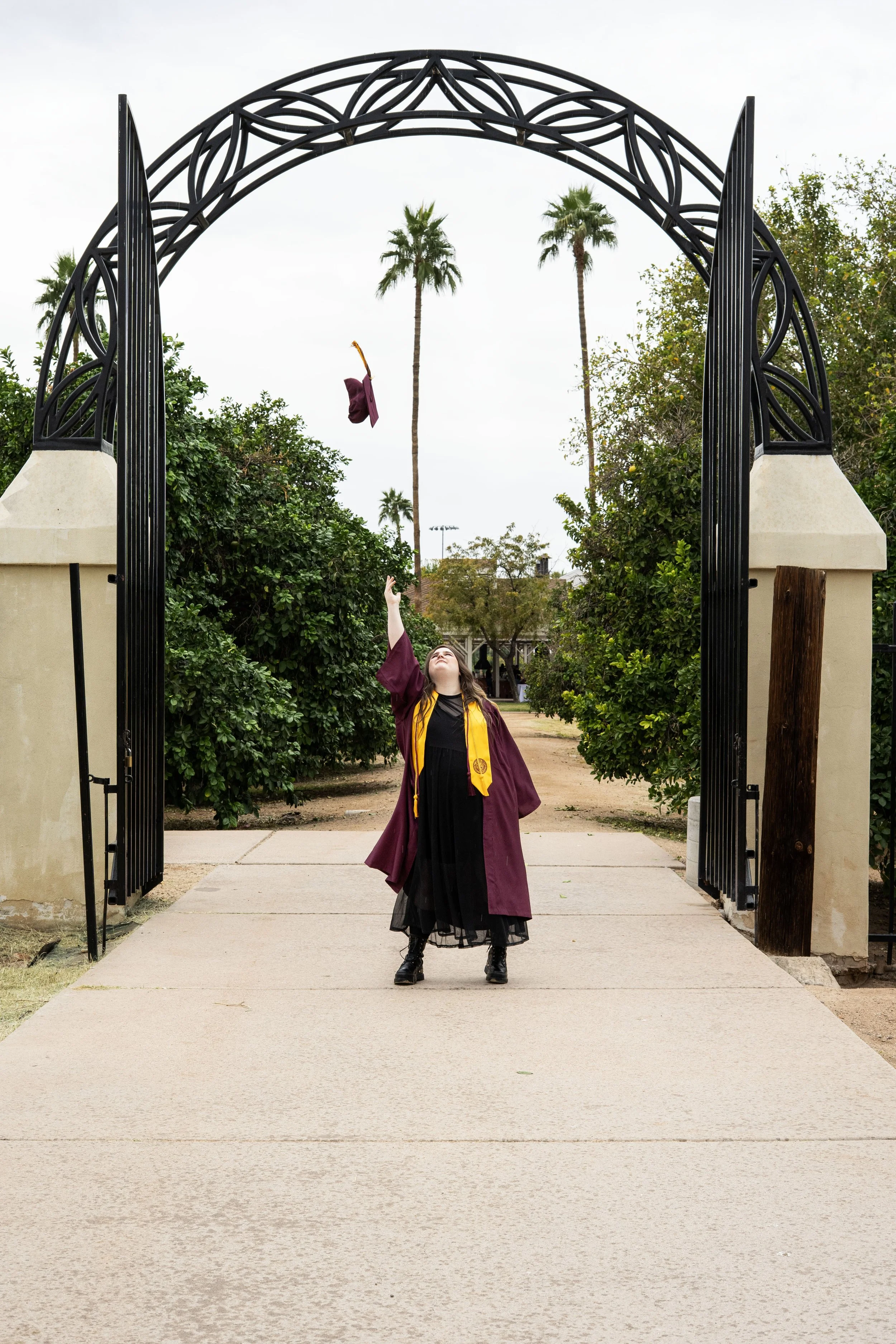 A young woman in graduation regalia stands on a sidewalk and throws her mortarboard cap into the air beneath a decorative archway, with palm trees and greenery in the background.