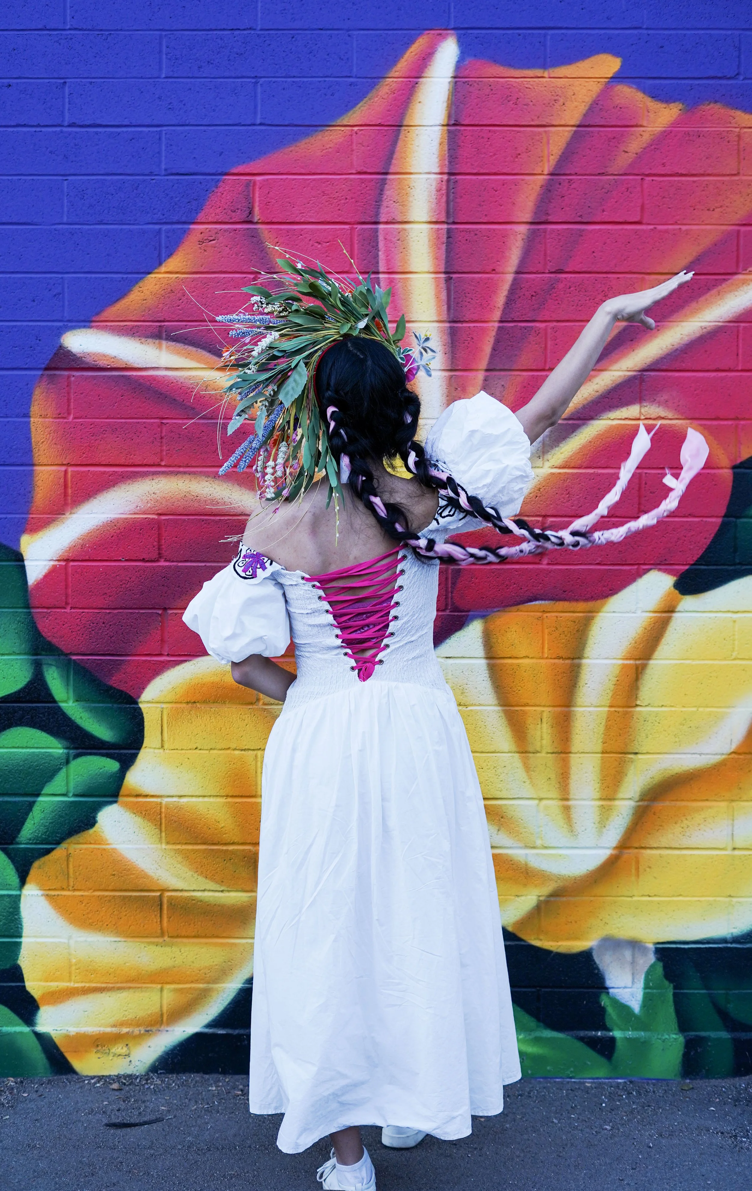 A woman dressed in a white dress with pink lacing on the back, wearing a large green leafy headpiece, stands in front of a colorful floral mural painted on a brick wall.