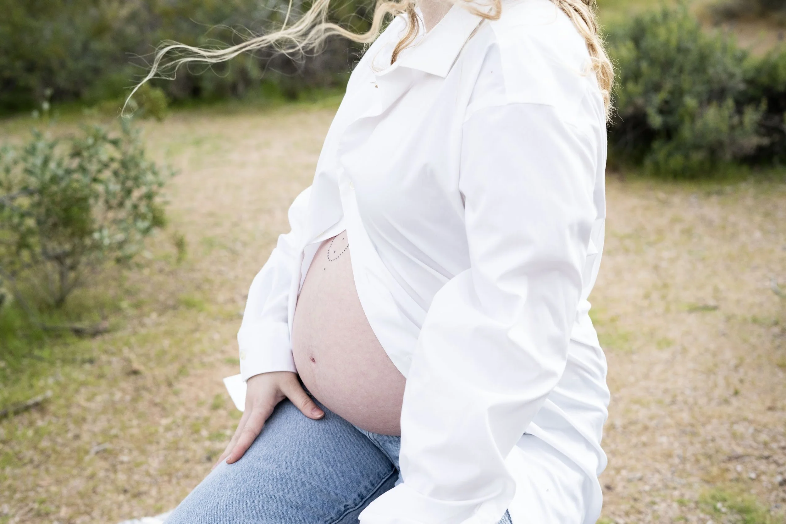 A pregnant woman with a visible baby bump, wearing a white shirt and jeans, sitting outdoors on a dirt path with greenery in the background.