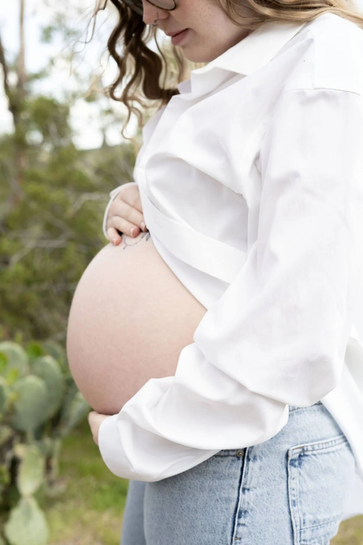 A pregnant woman with long, curly hair, wearing glasses, a white shirt, and jeans, standing outdoors in a natural setting with greenery and cactus plants.