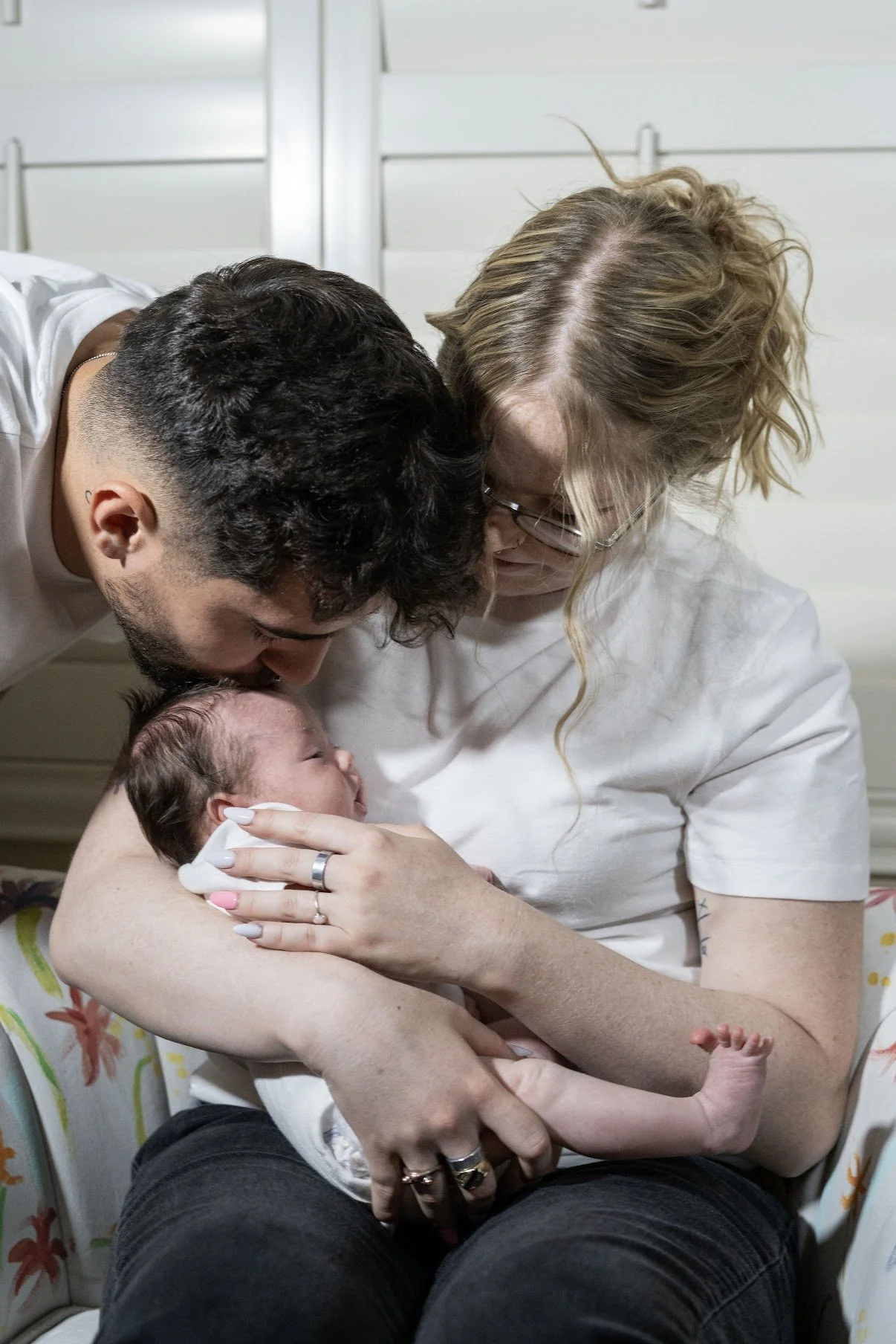 A woman holding a newborn baby with a man leaning in, both touching the baby's head, in a hospital room.