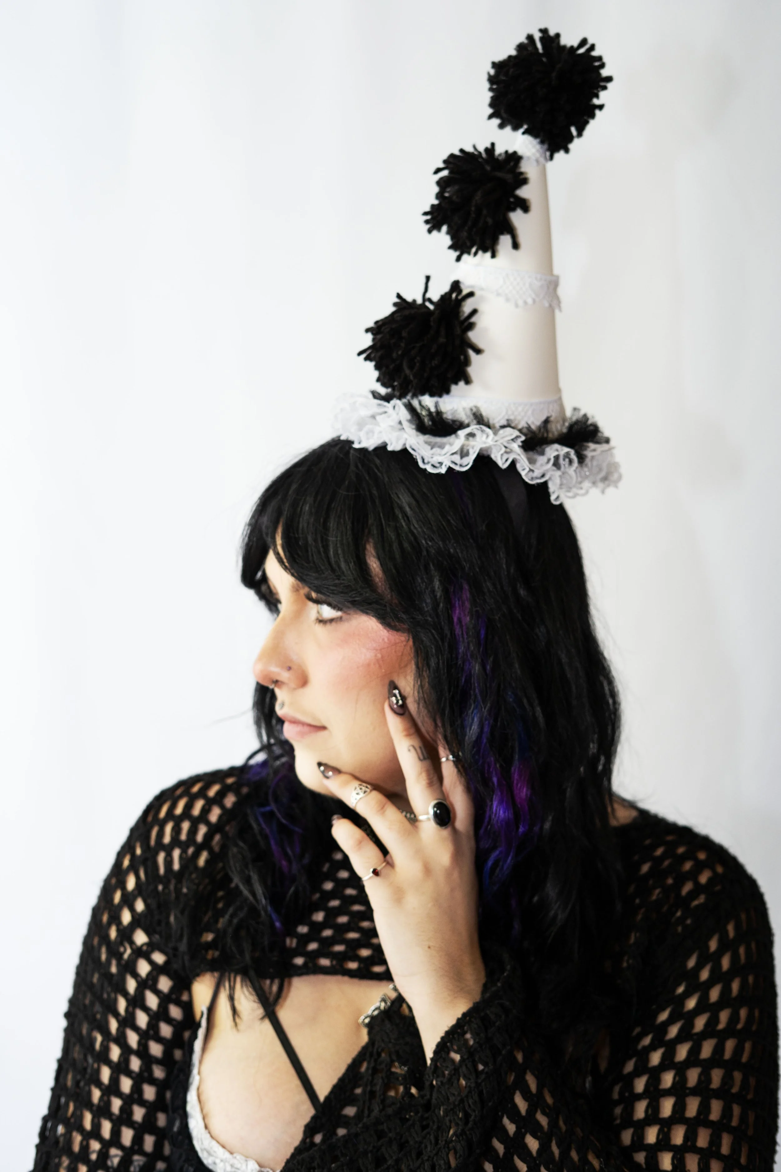 A woman with black hair and purple streaks wearing a black fishnet top. She has piercings, rings, and tattoos, and is touching her face with her right hand. She is wearing a tall, whimsical white hat with black pom-poms and lace decorations.