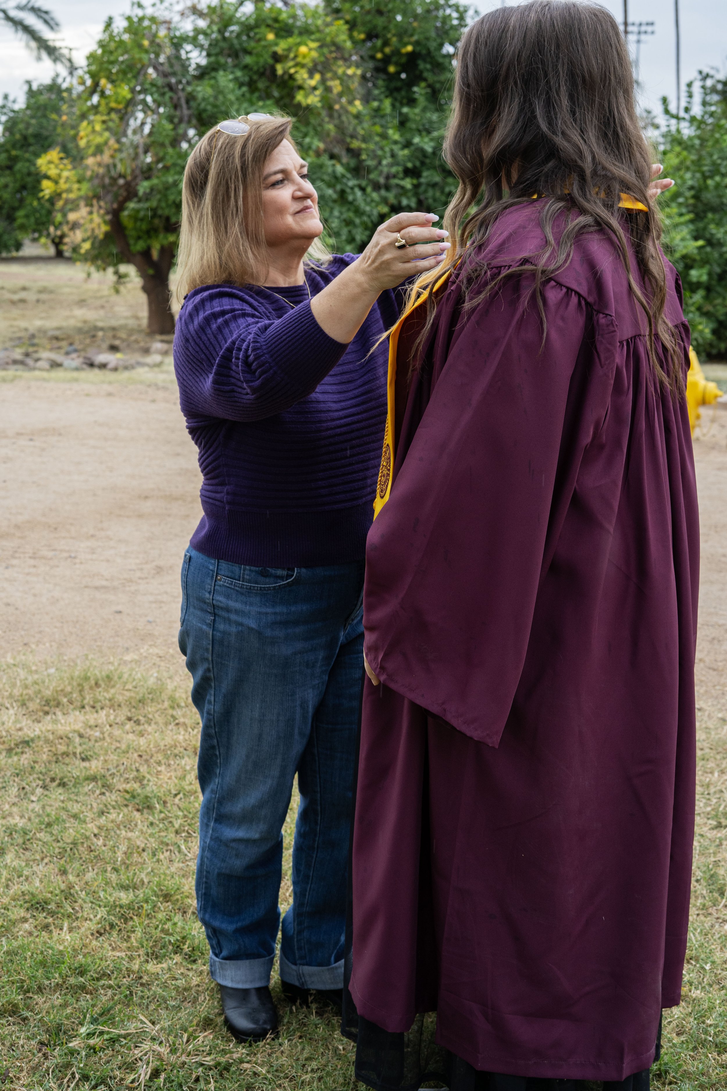 A woman in a purple sweater is giving a medal to a graduate wearing a maroon cap and gown outdoors in a park with trees and grass.
