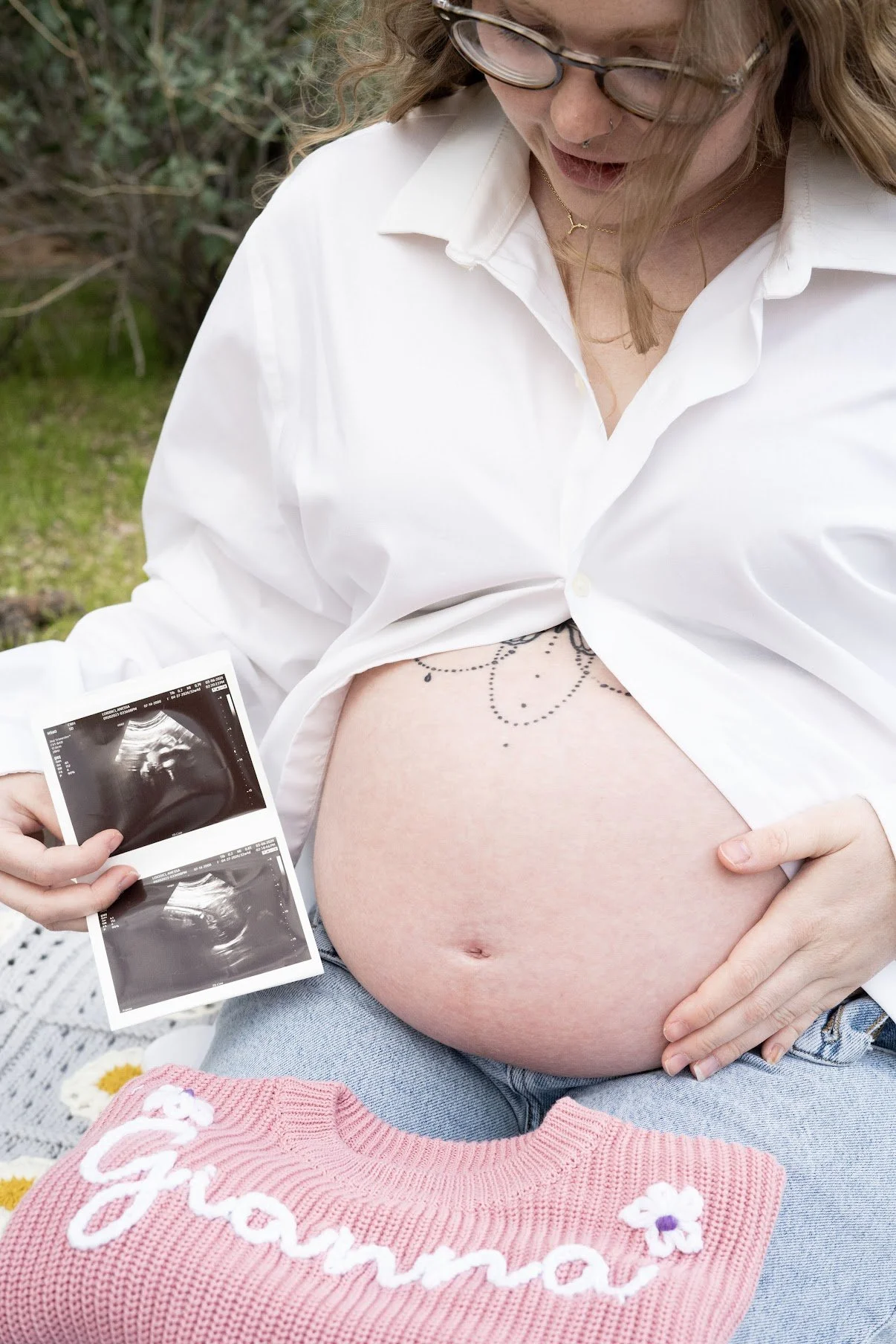 Pregnant woman holding ultrasound images and revealing her belly with decorative tattoo, wearing a white shirt and blue jeans, sitting outdoors on a blanket with a pink knit sweater reading 'Glamma' nearby.