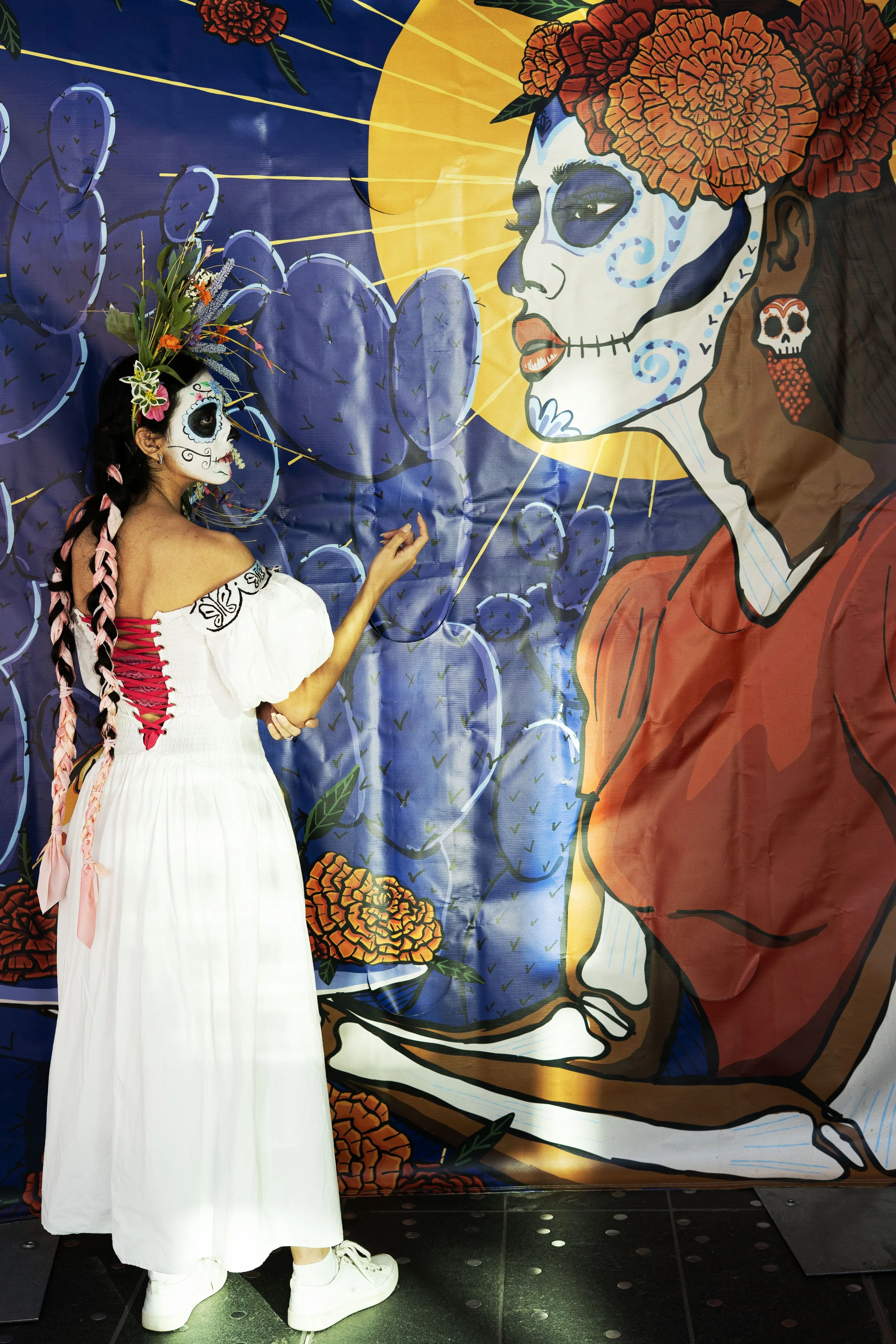 A woman dressed in a white dress with red accents and a sugar skull face paint, standing in front of a colorful mural showcasing a woman with sugar skull makeup, marigold flowers in her hair, and a large yellow moon in the background.