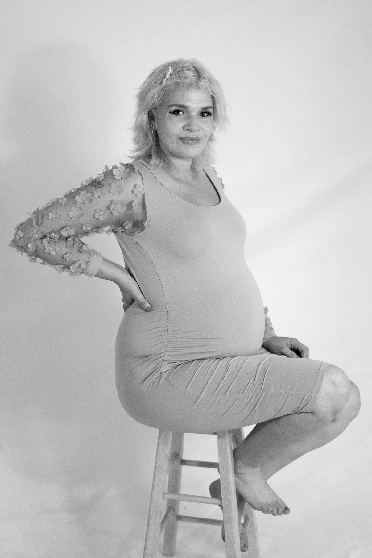 A pregnant woman sitting on a wooden stool, posing with one hand on her hip, smiling softly, wearing a long dress with floral sleeves, in a studio with a plain background.
