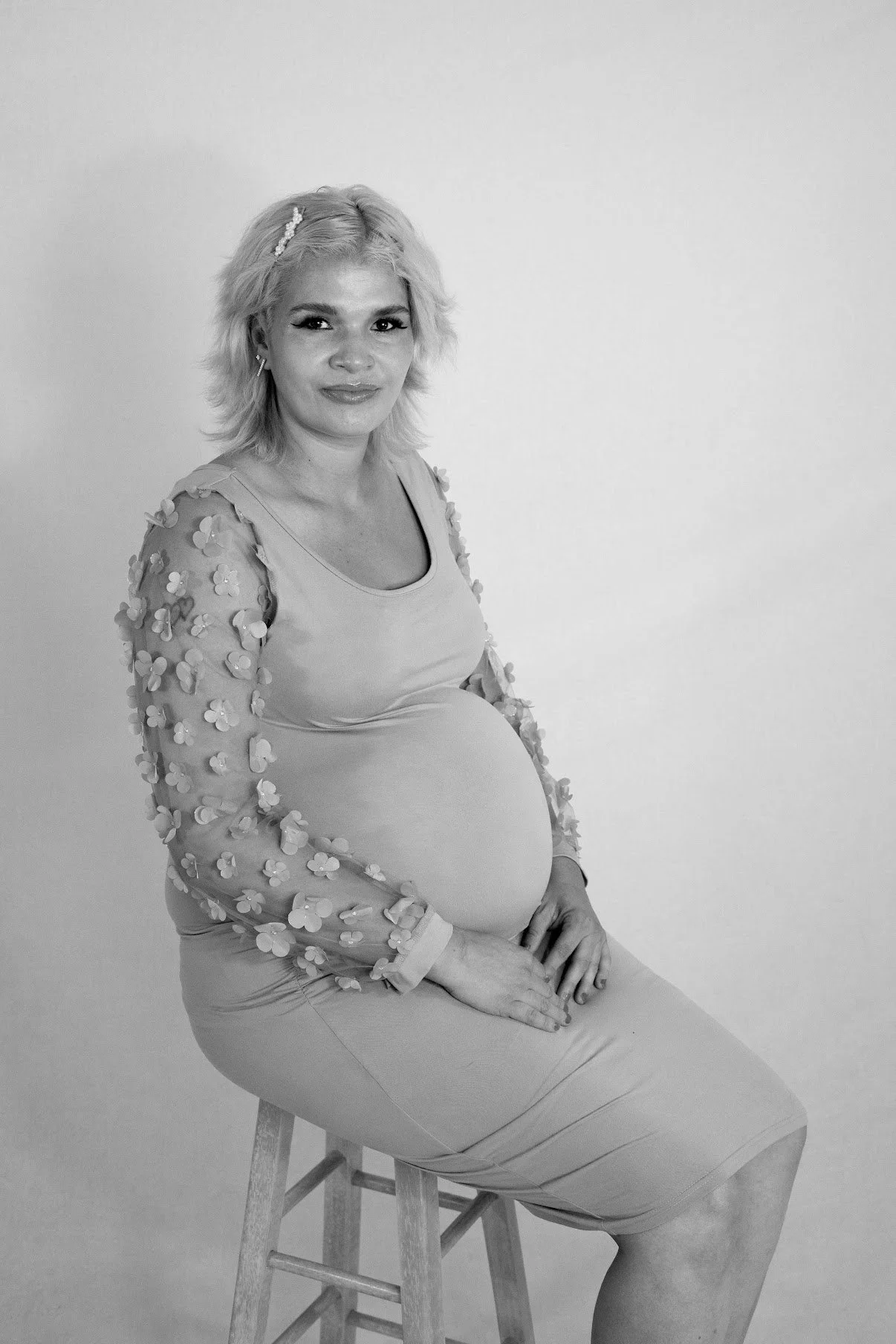 A pregnant woman with light-colored, shoulder-length hair sitting on a stool against a plain wall. She is wearing a dress with sheer, floral-embellished sleeves and is looking at the camera with a gentle expression.