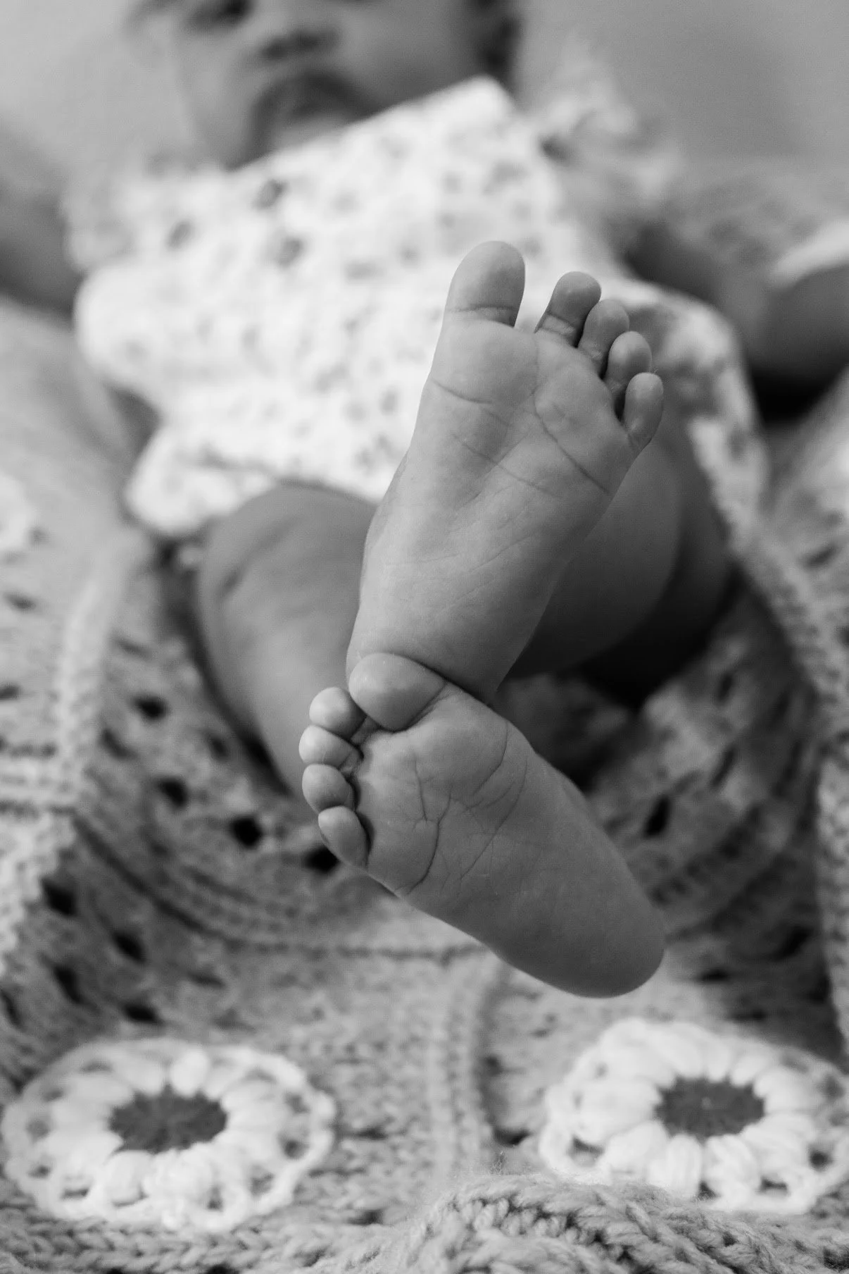 Close-up of a newborn baby's feet with toes curled up, laying on a crocheted blanket, with a blurred face in the background.