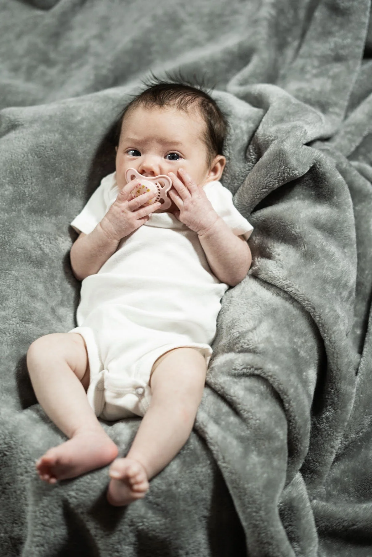 A baby lying on a gray blanket, holding a pacifier to their mouth, and looking at the camera.