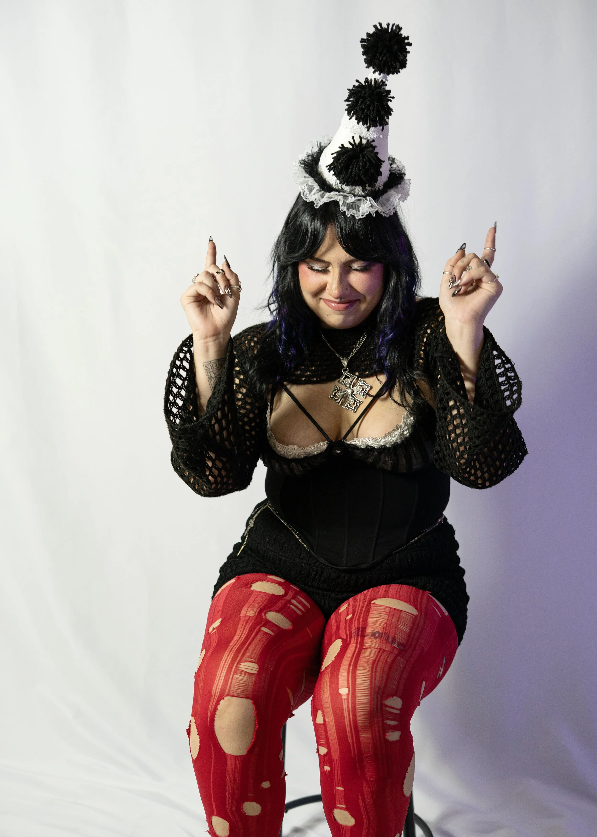 A woman with black hair, wearing a black crochet-style top, a black corset, red torn stockings, and a festive party hat with pom-poms, standing against a plain white background.