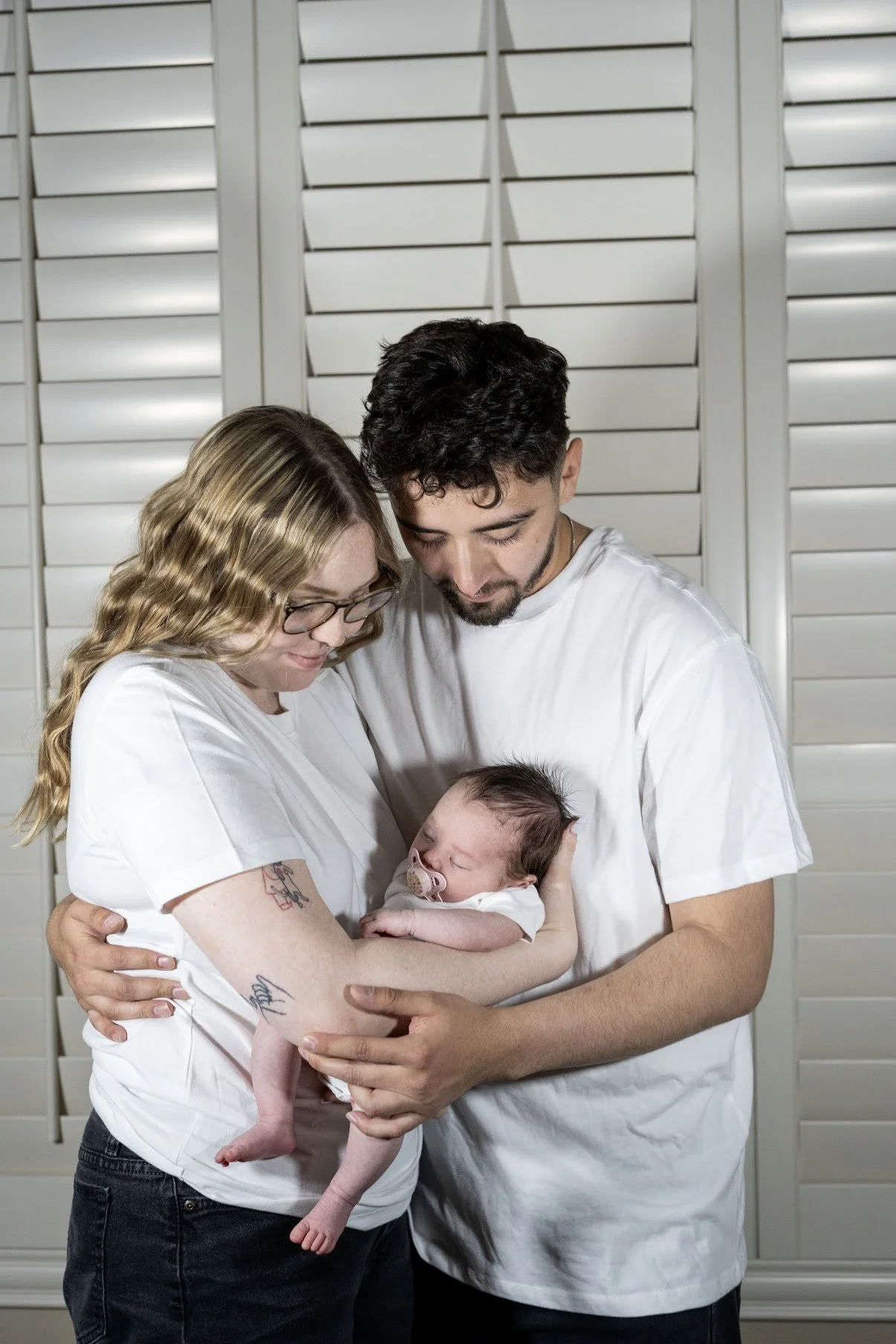 A couple holding their newborn baby in a bright room with white shutters.