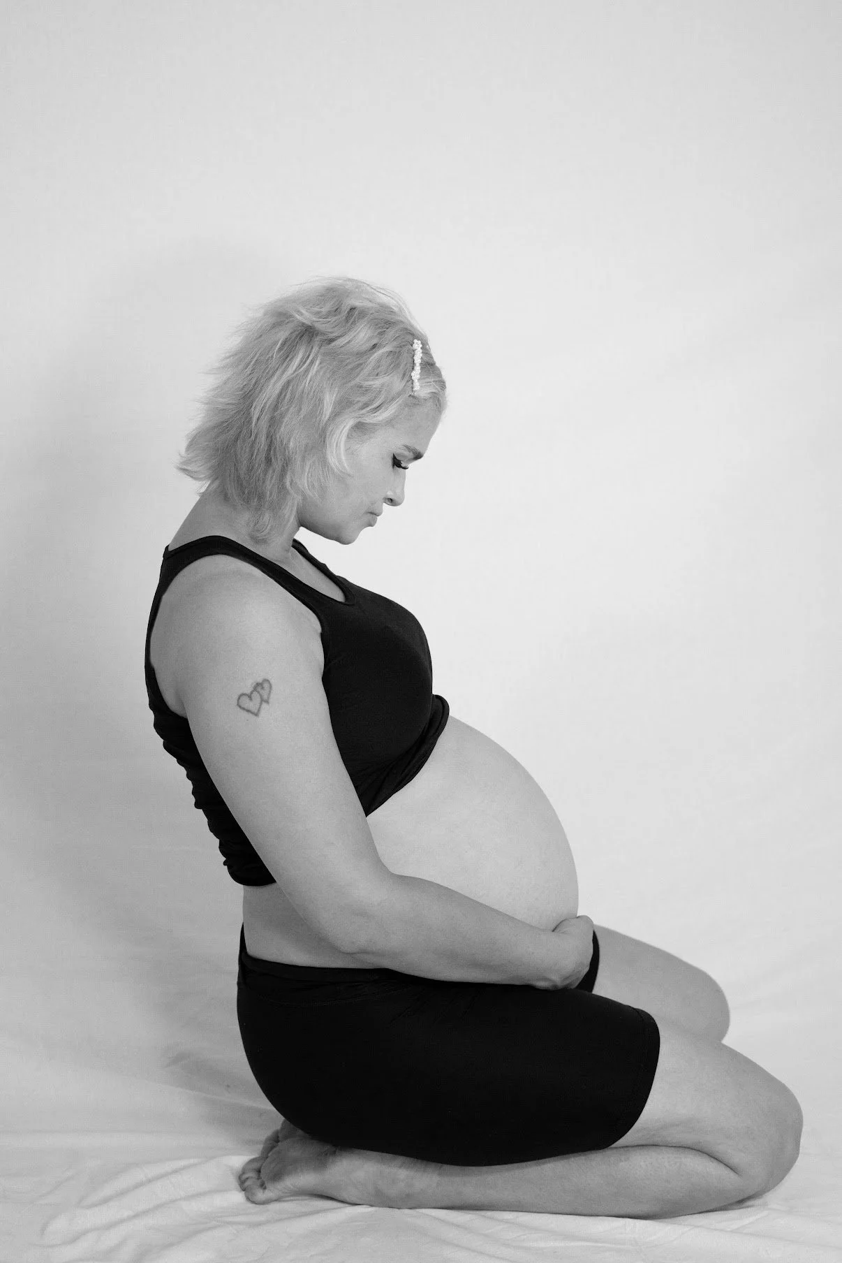 A pregnant woman with short blonde hair sitting on the floor, knees drawn up, with her head bowed and eyes closed, wearing a black sleeveless top and black shorts, against a plain white background.