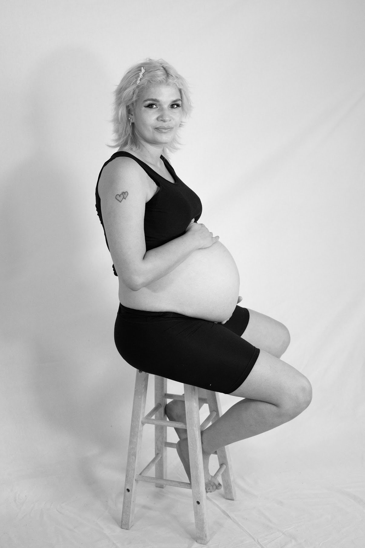 Black and white photo of a pregnant woman sitting on a wooden stool, wearing a sleeveless top and shorts, with her hands on her belly, and looking at the camera.