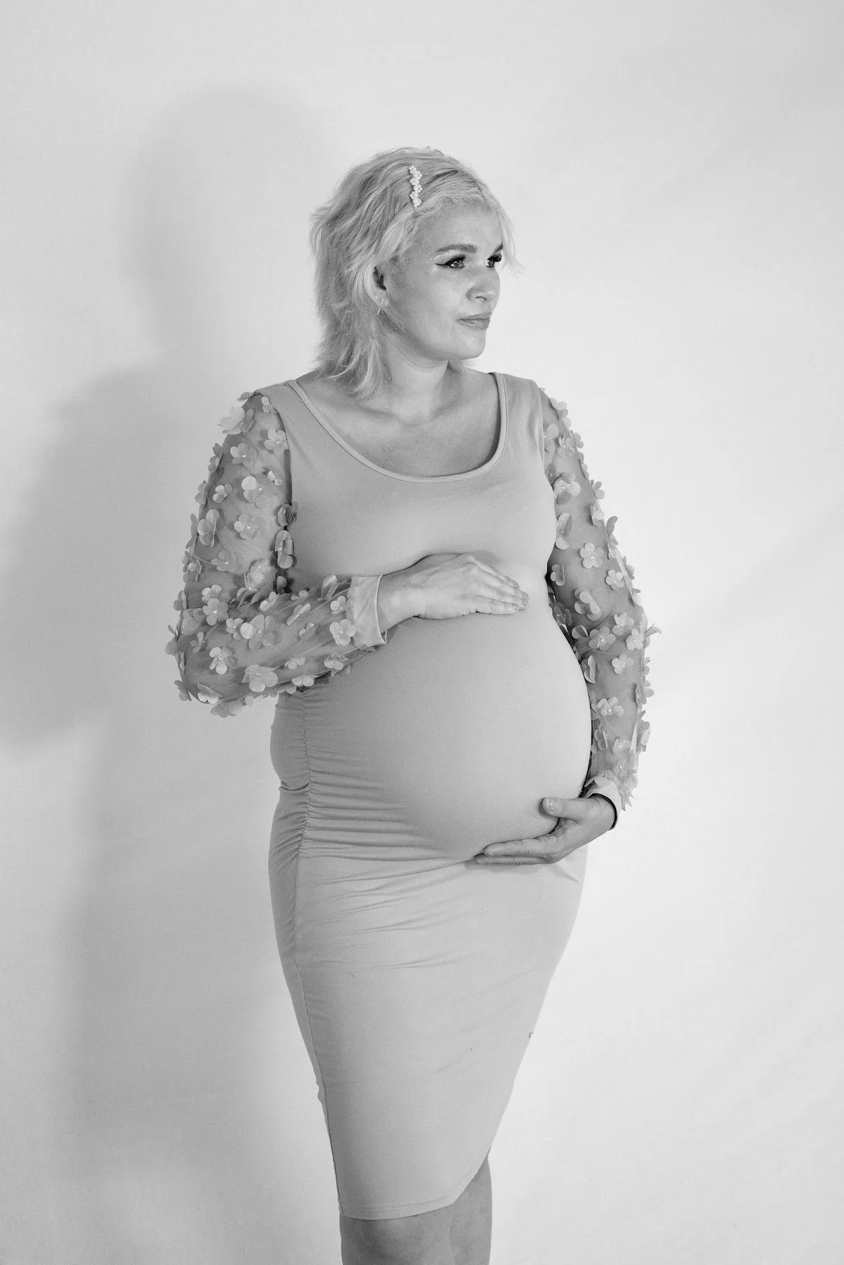 A pregnant woman standing in front of a plain wall, holding her belly with one hand, wearing a fancy dress with floral, transparent sleeves.
