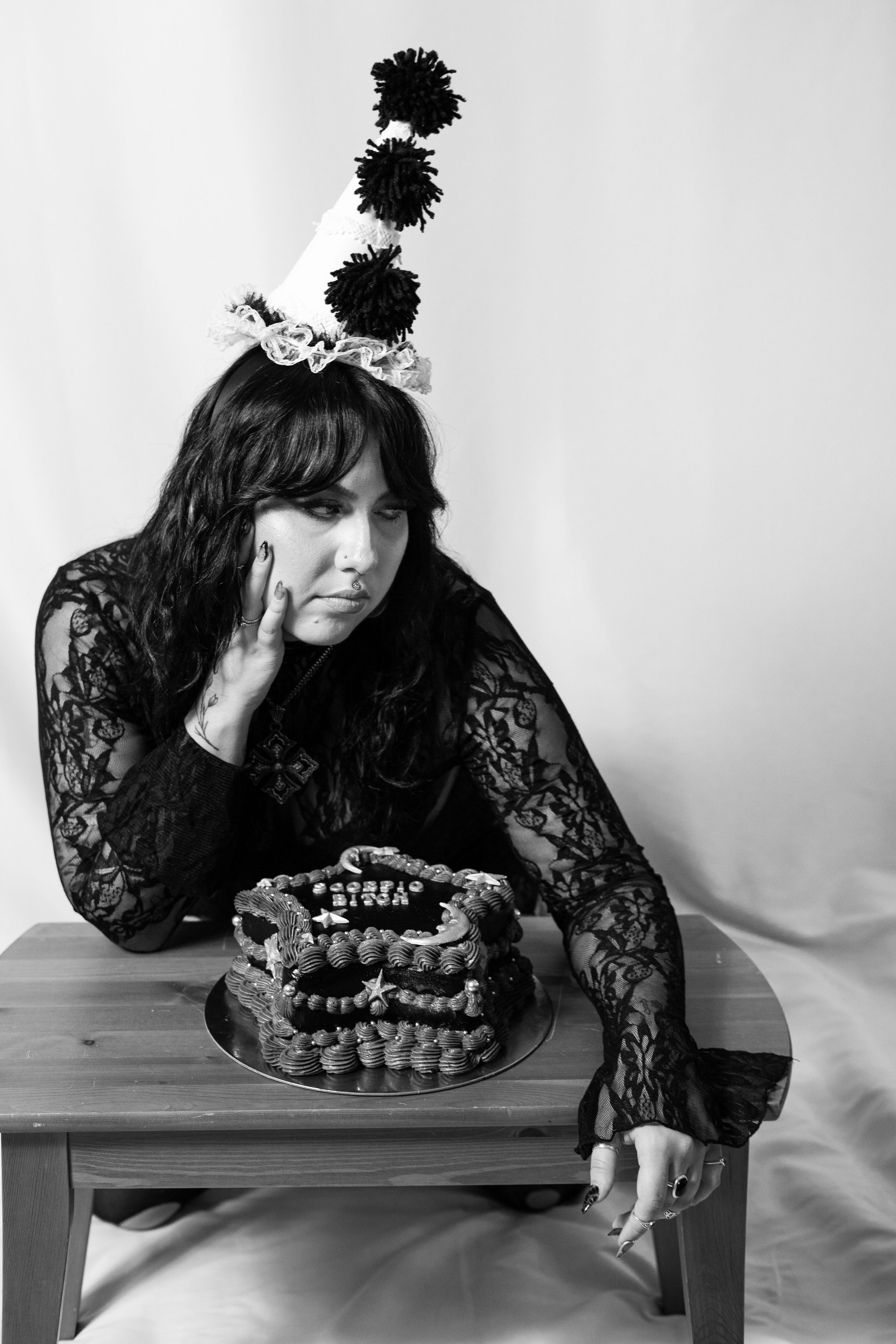 A woman wearing a ruffled party hat sitting at a table with a cake, her hand on her face, looking disappointed.