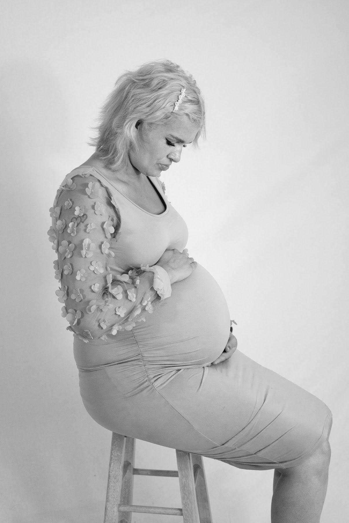 A pregnant woman with wavy blonde hair sits on a wooden stool with her eyes closed, gently holding her belly, wearing a light-colored top with 3D floral embellishments on the sleeves and a fitted skirt, against a plain background.