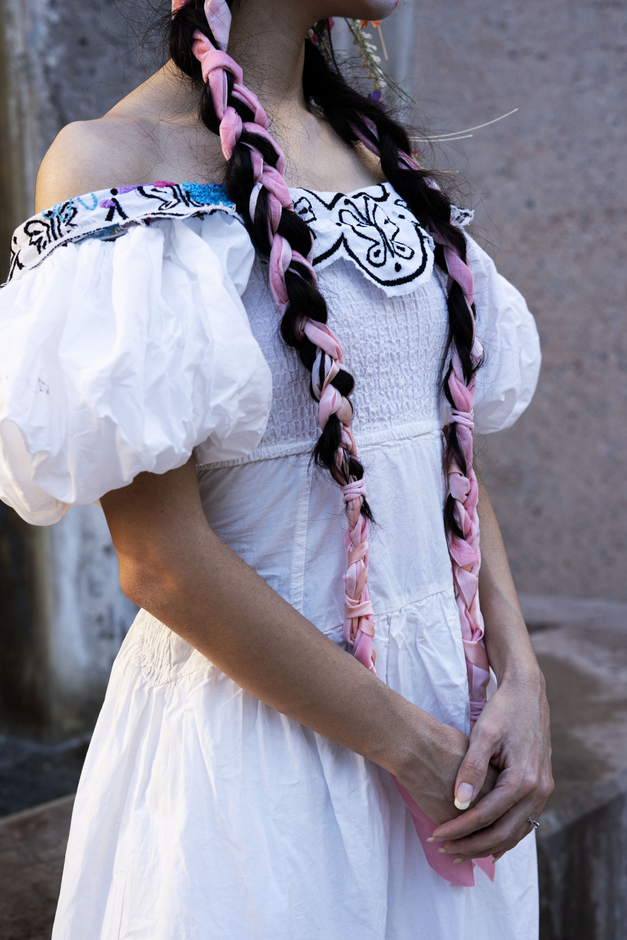 A woman wearing a traditional white dress with embroidered details and puffed sleeves, with long braided hair in pink and black.