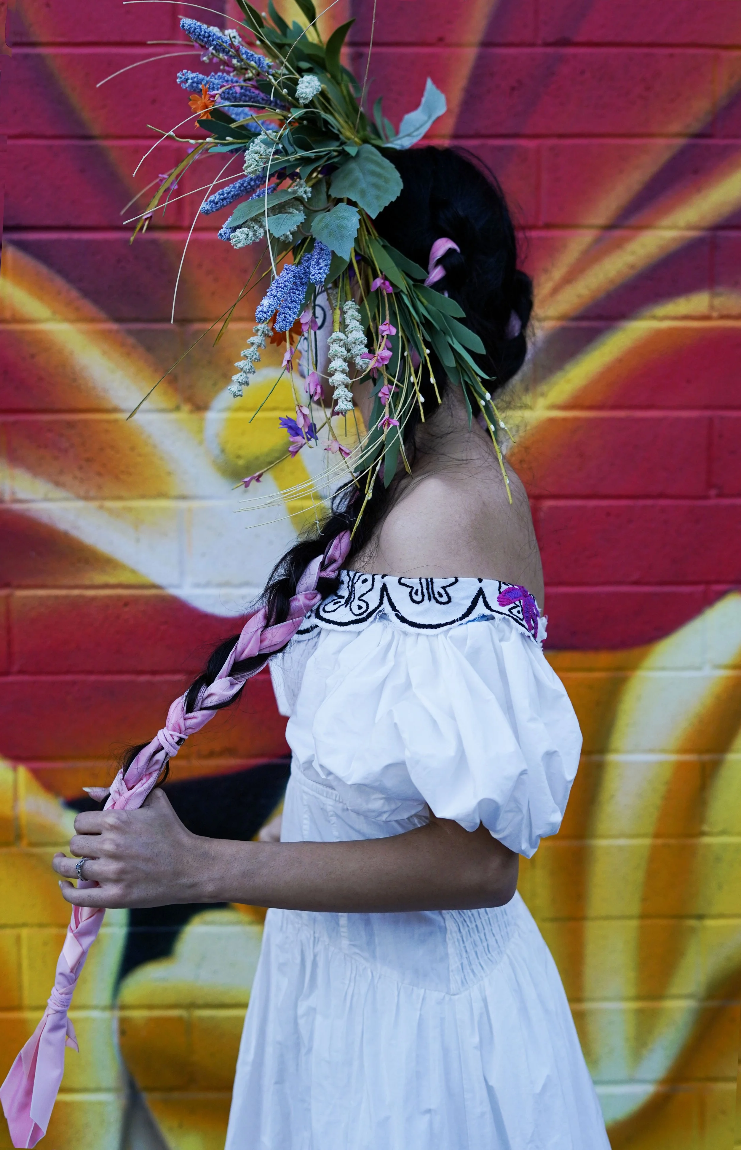 A woman with a long braid, dressed in a white off-the-shoulder dress with embroidery, stands in front of a colorful mural holding a large bouquet of flowers on her head.