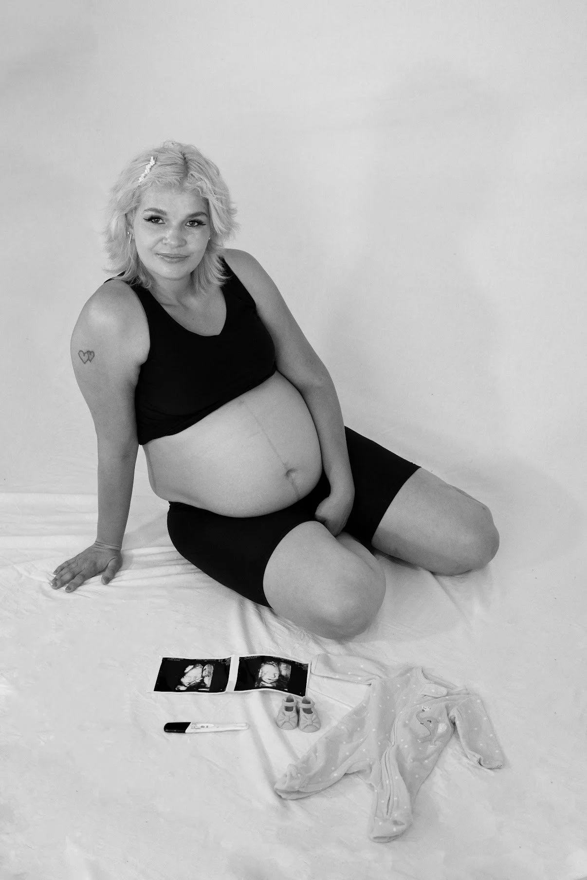 A pregnant woman with blonde hair sitting on the floor, wearing a black tank top and black shorts, smiling at the camera. In front of her, there are ultrasound photos, a small pair of baby shoes, a pregnancy test, and baby clothing.