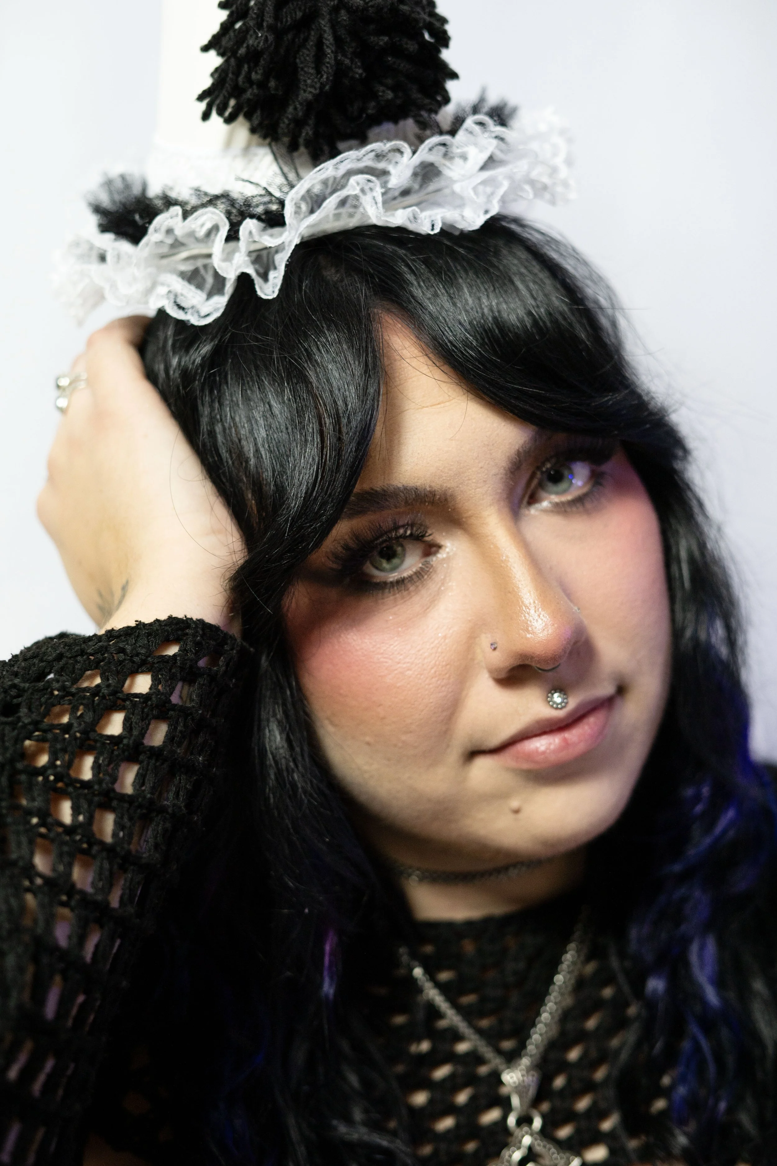 A young woman with black hair and light skin, wearing dark eye makeup and a nose piercing, dressed in a black mesh top with jewelry, is posing with her hand resting on her head, and is wearing a decorative headpiece with black and white ruffles.
