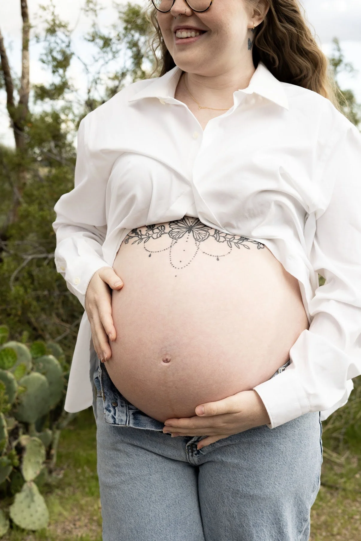 A pregnant woman standing outdoors, smiling, wearing a white shirt and jeans, with floral tattoo above her belly and holding her belly with both hands.