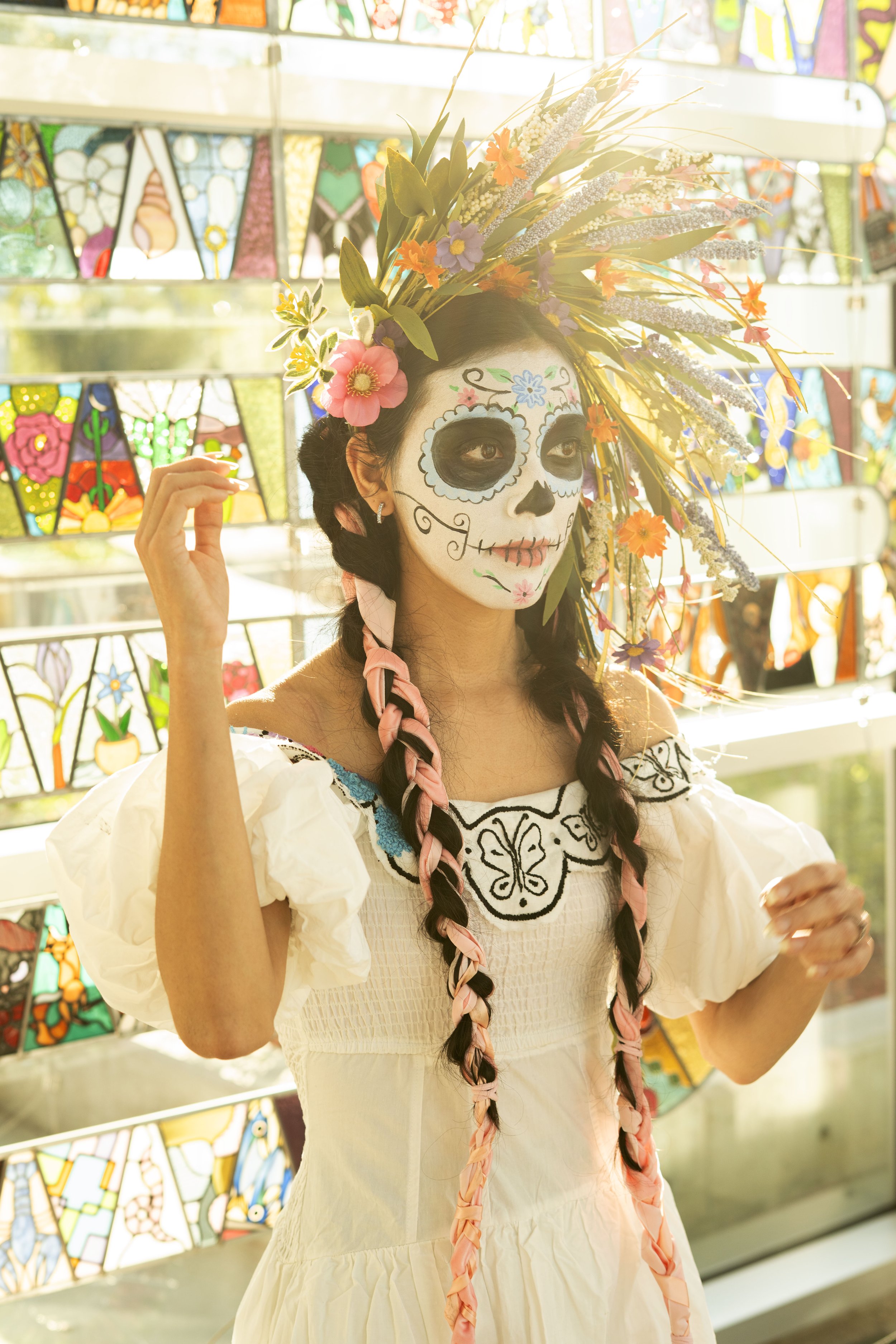 Woman dressed in colorful traditional attire with a sugar skull face paint and a large floral headdress, standing in front of a stained glass display.