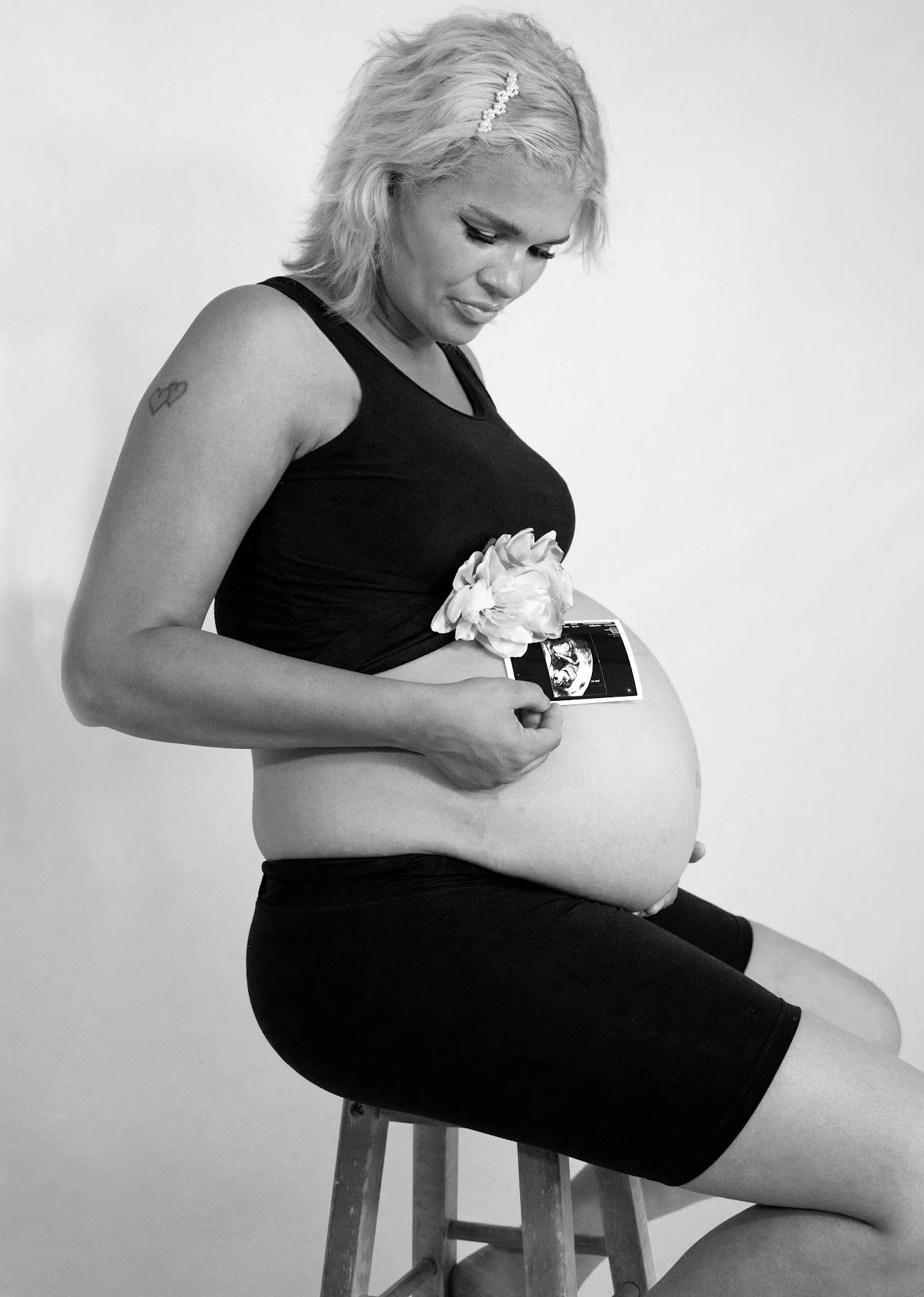 Black and white photo of a pregnant woman sitting on a stool, holding an ultrasound picture on her belly, with a flower decoration on her clothing, and looking down at her belly.