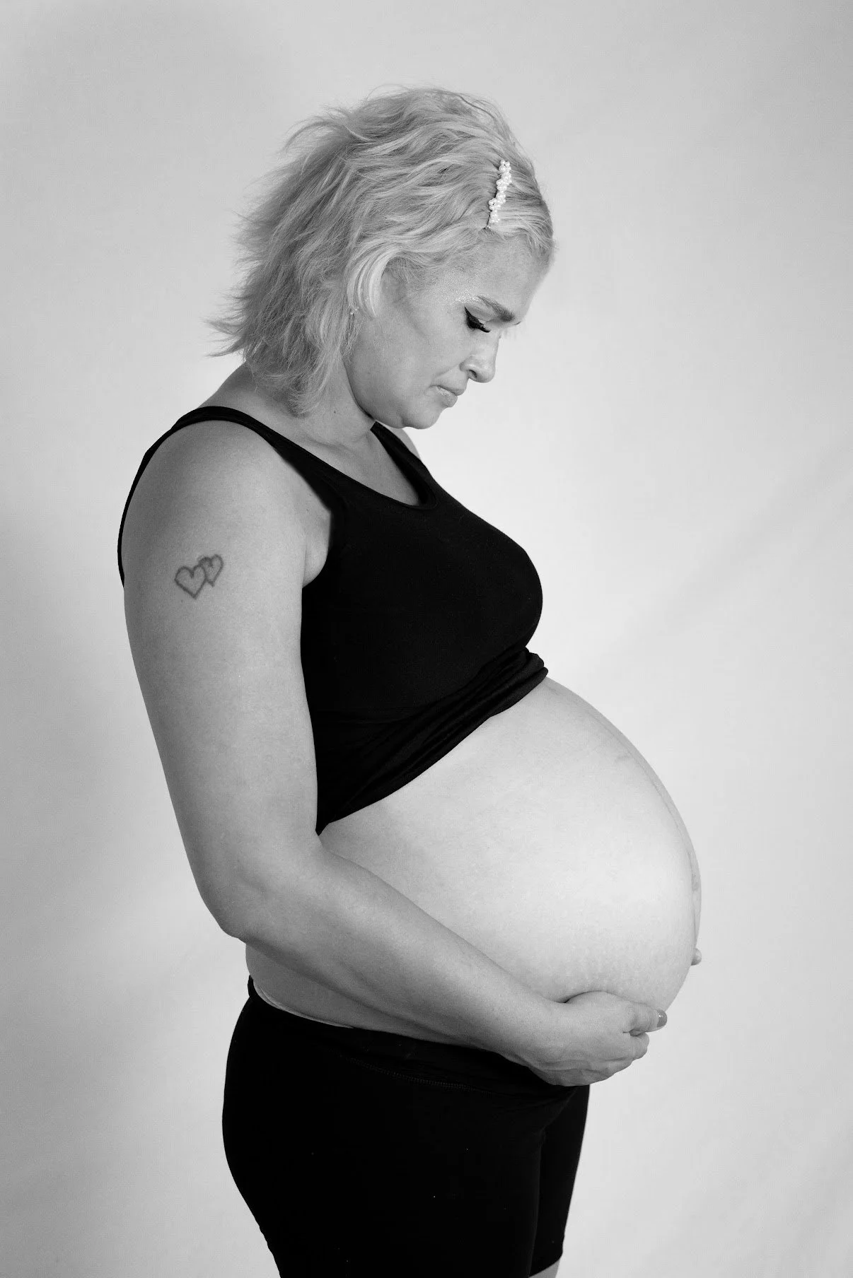Black and white photo of a pregnant woman with blonde hair and a tattoo of two connected hearts on her arm, wearing a black tank top and black pants, standing against a plain background, with her hands resting on her belly, looking down.