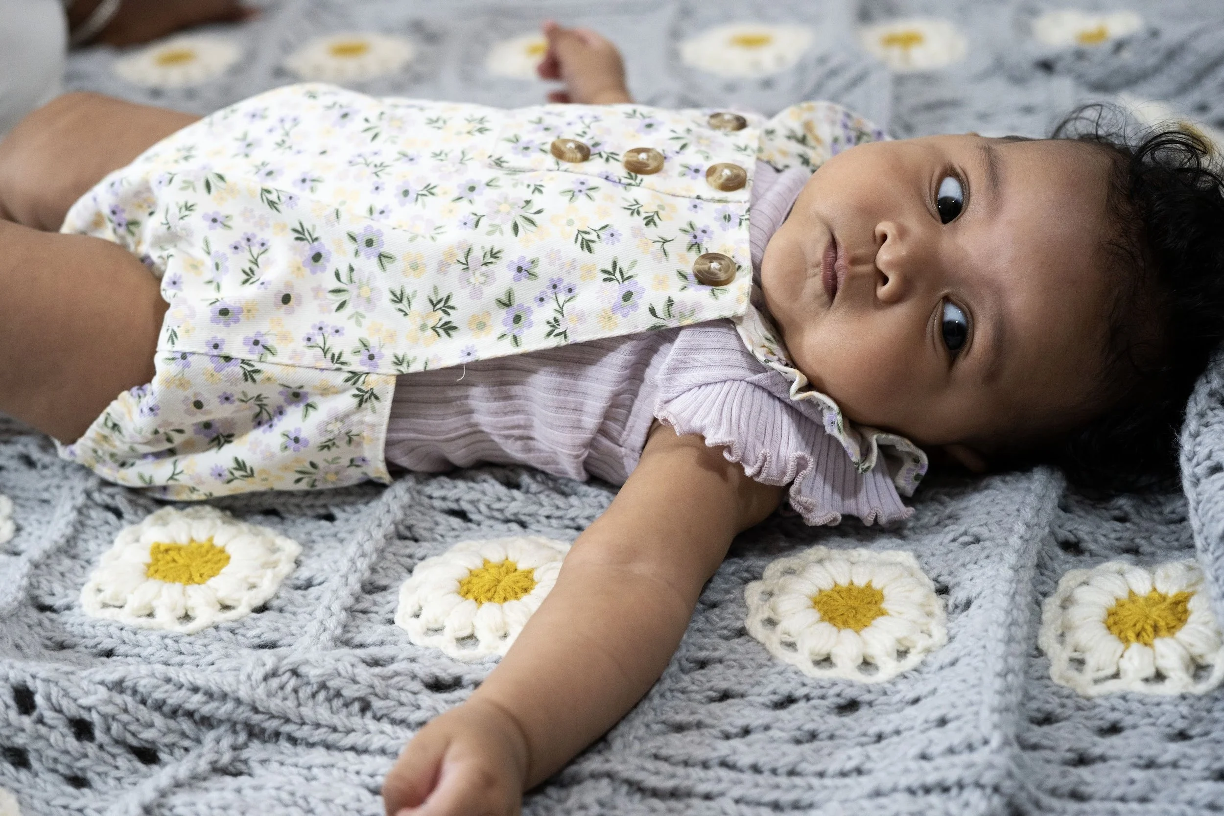 Baby lying on a crocheted blanket with yellow and white flower patterns, wearing a floral dress with buttons, gazing towards the camera.