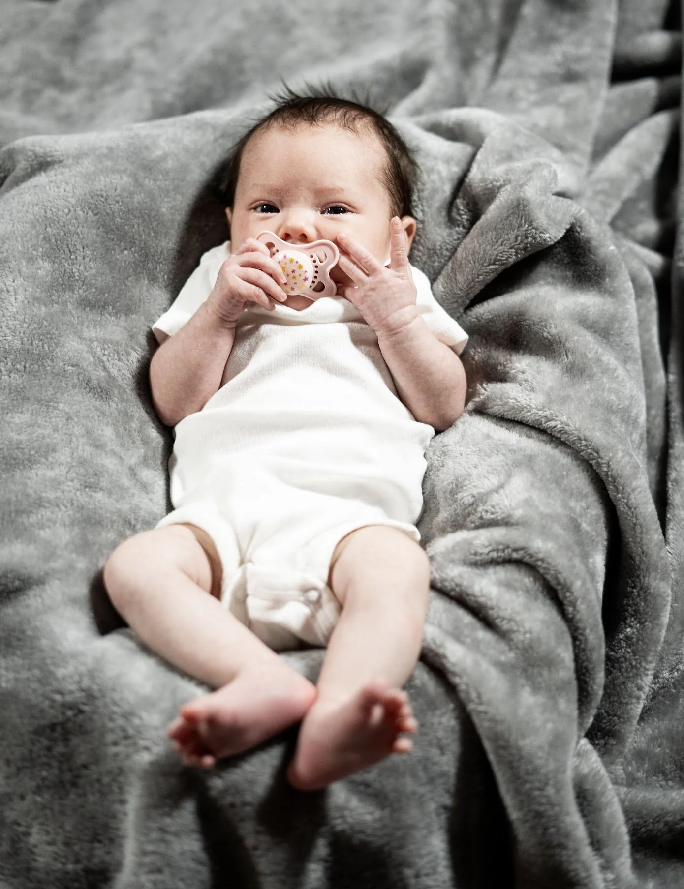 An infant lying on a gray blanket, holding a pacifier, wearing a white onesie.
